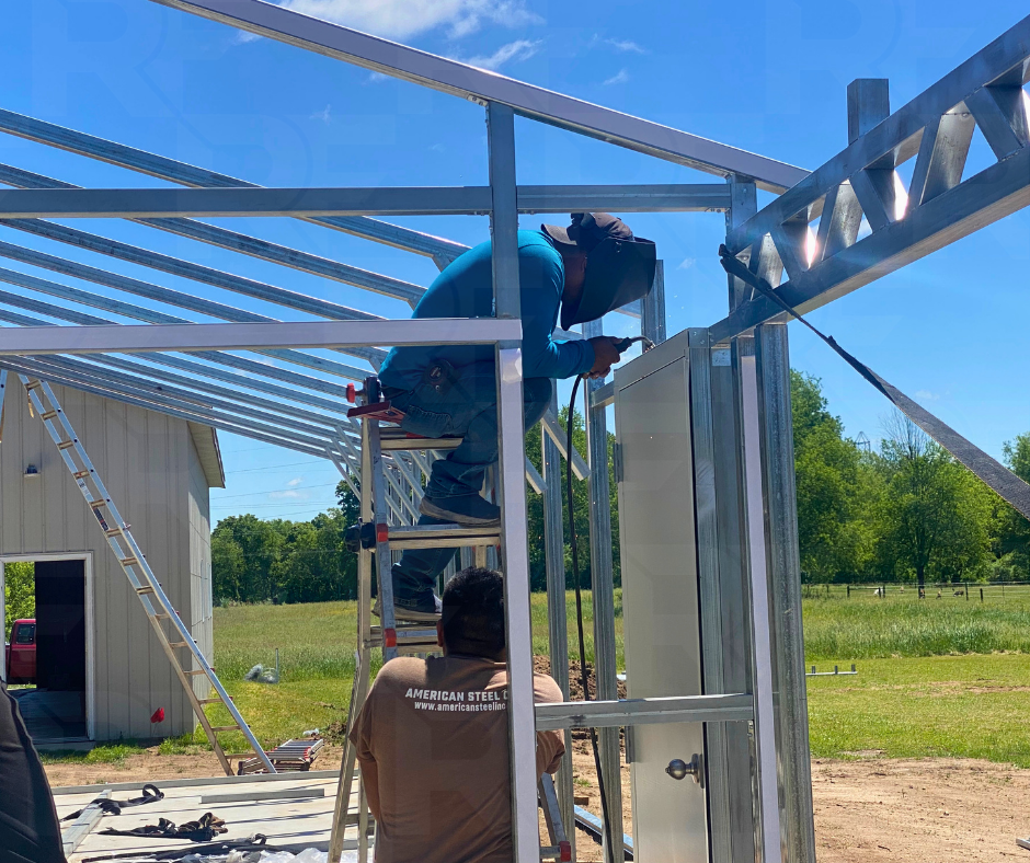 Two workers assembling a metal structure outdoors under a clear blue sky, with one standing on a ladder and using a power tool, while the other stands below observing.