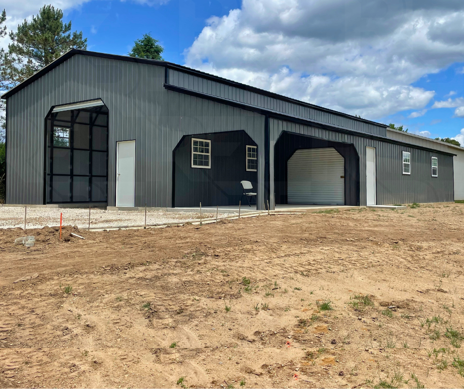 A large metal building with black metal exterior, multiple small windows, and a roll-up garage door, situated on a dirt lot under a partly cloudy sky.