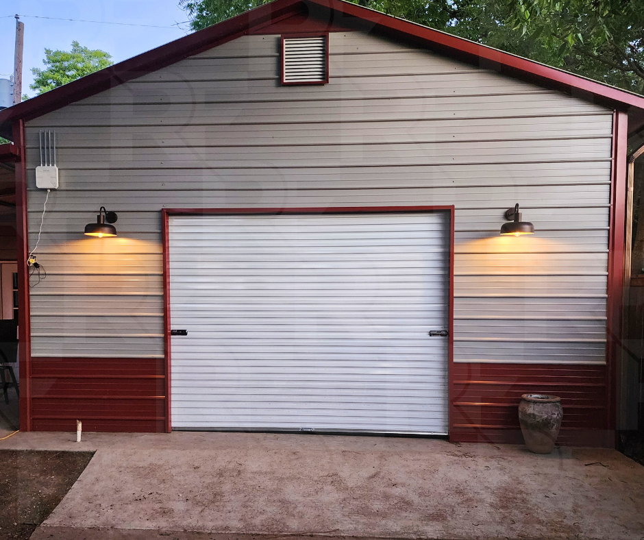 A shed or garage with gray and red siding, illuminated by two wall-mounted exterior lights, with a large white roll-up door, a small ventilation grate at the top, and a potted plant to the right.