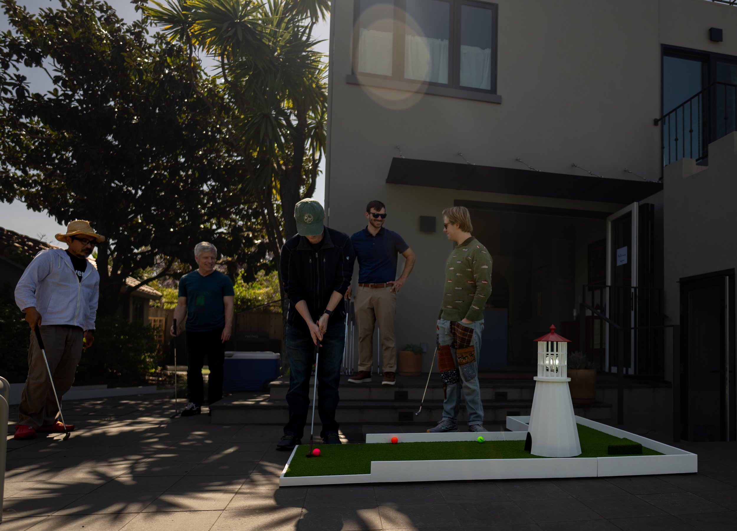 Group of five men playing miniature golf outdoors on a patio in front of a modern house, with one man taking a shot, a small lighthouse decoration, and trees in the background.