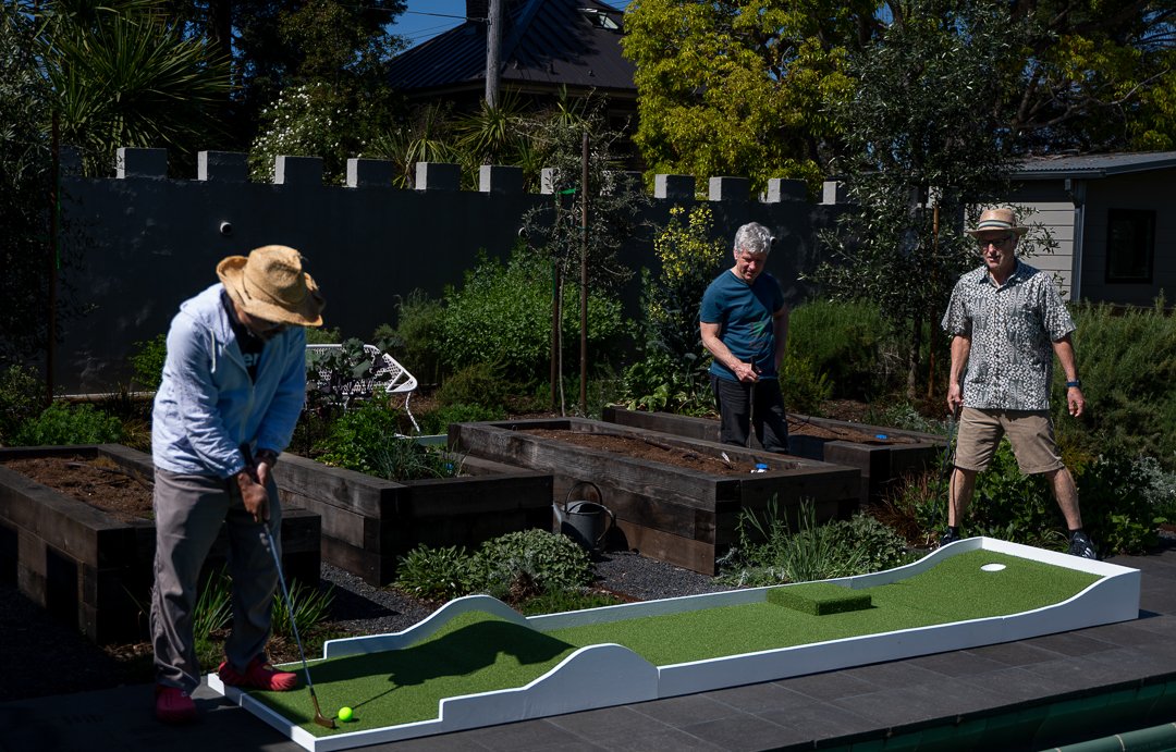 Three men playing miniature golf in a backyard garden with raised garden beds and trees.