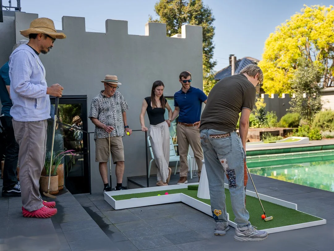 Group of people playing mini golf by a pool on a sunny day, some watching and some swinging golf clubs.