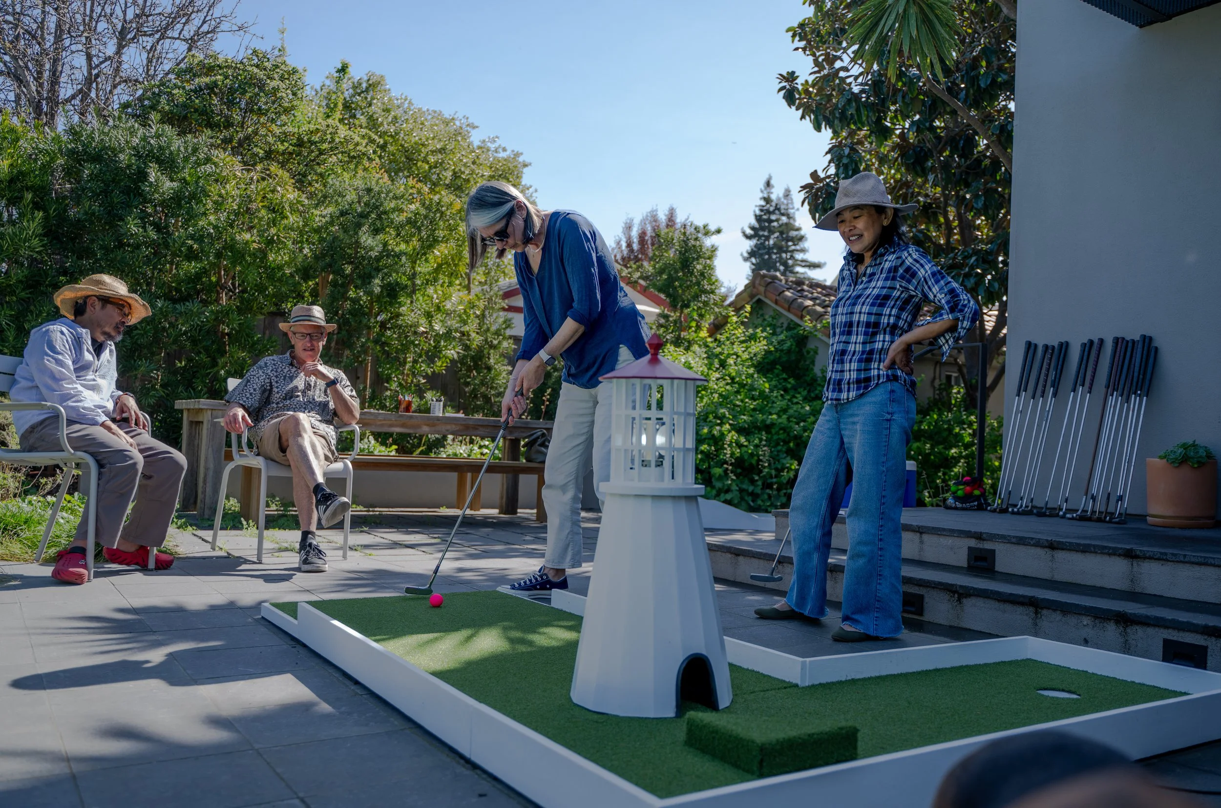 A group of four people playing mini golf outside on a sunny day. Two women are standing on the mini golf course, preparing to putt, while two men are sitting on chairs nearby watching. The scene is set on a patio with trees and plants in the backgrou