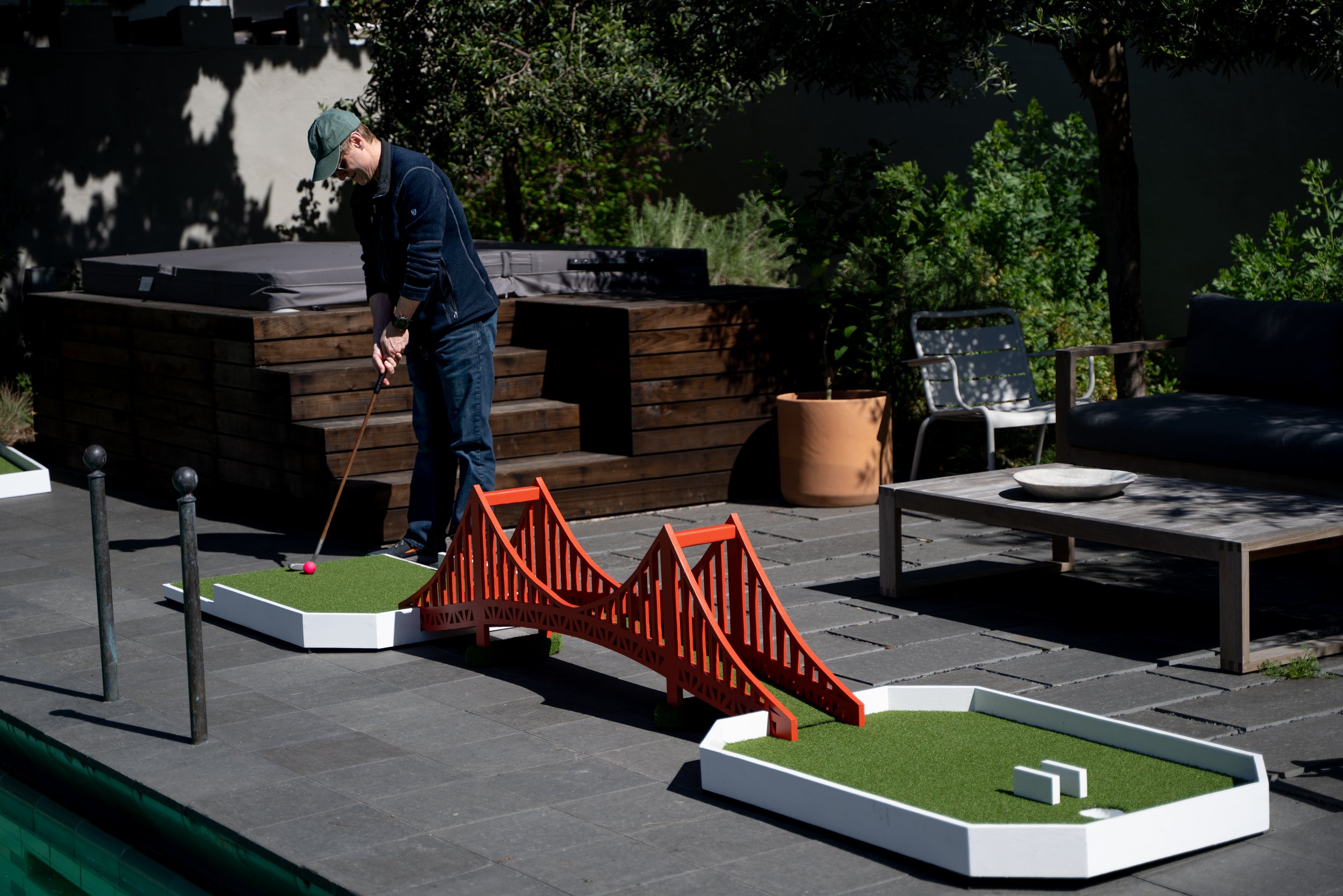 A man playing mini golf outdoors on a paved patio, with a miniature bridge and cityscape backdrop.