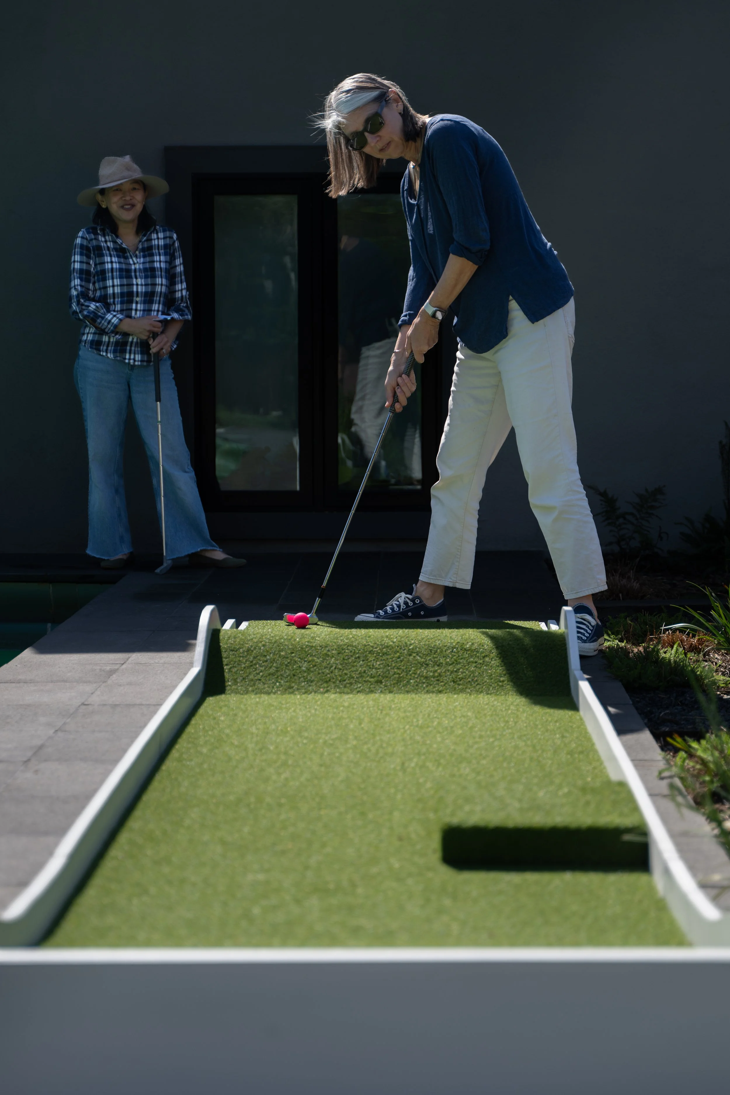 Two women playing mini golf outdoors, one preparing to hit the golf ball while another watches.