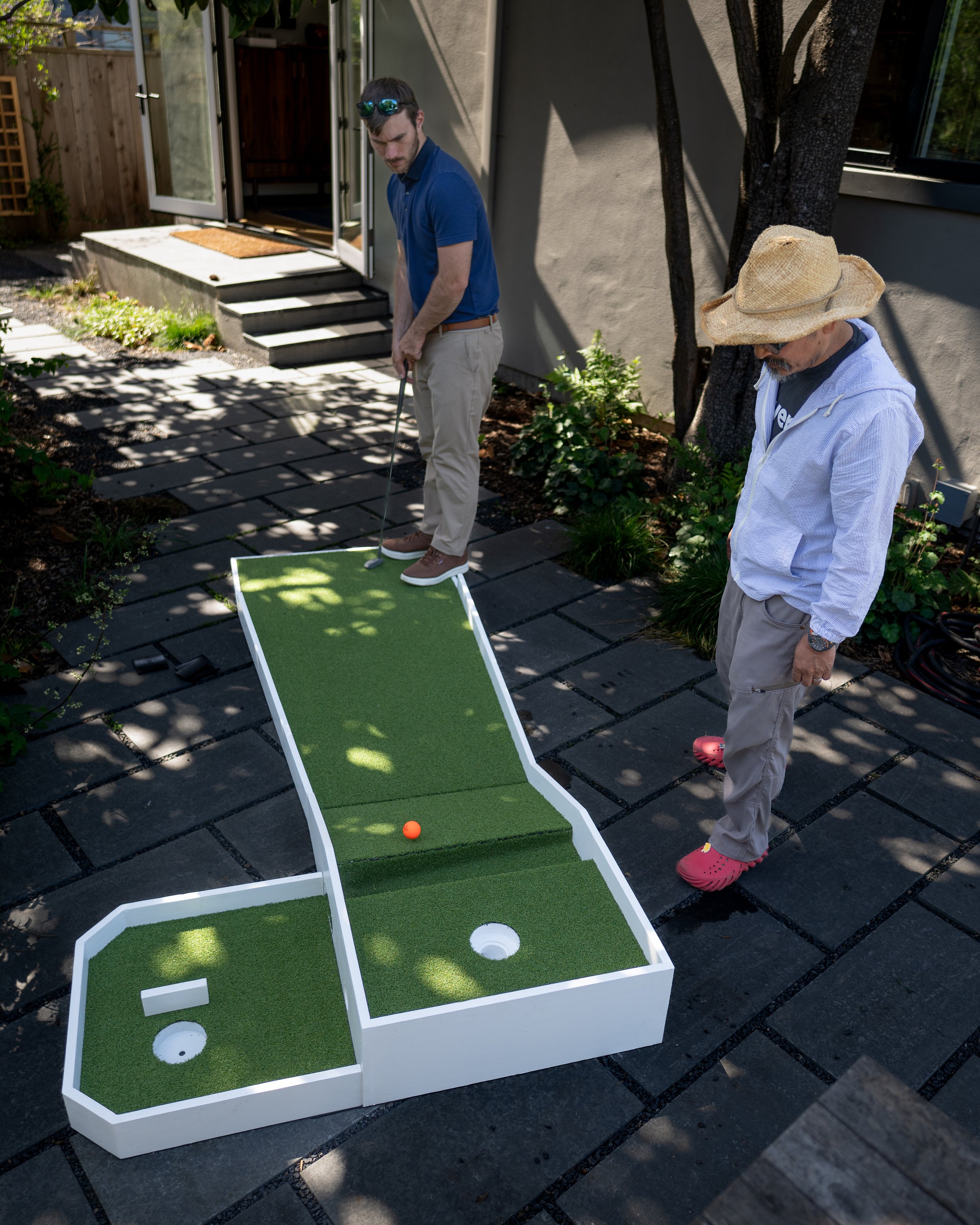 Two men are playing mini golf outdoors on a sunny day. One man is about to hit a golf ball, while the other watches. The mini golf course has a raised green with a hole and a ramp, placed on a stone patio near a garden with plants and a tree.