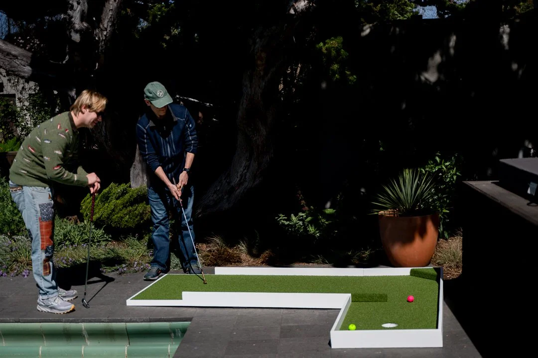 Two young men playing mini-golf outdoors on a sunny day, with a mini-golf course featuring a green turf in a small rectangular area, a pink golf ball, a green golf ball, and a potted plant nearby.