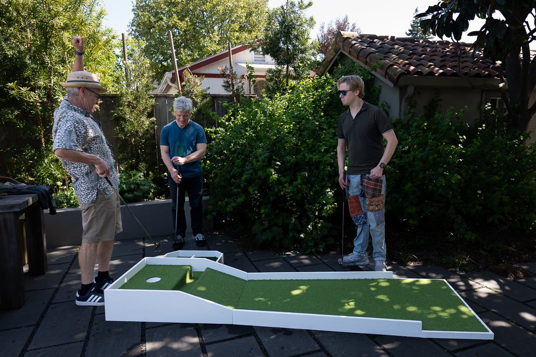 Three men playing miniature golf outdoors on a sunny day, with one man swinging a club, and a mini golf course with green artificial turf in front of them.