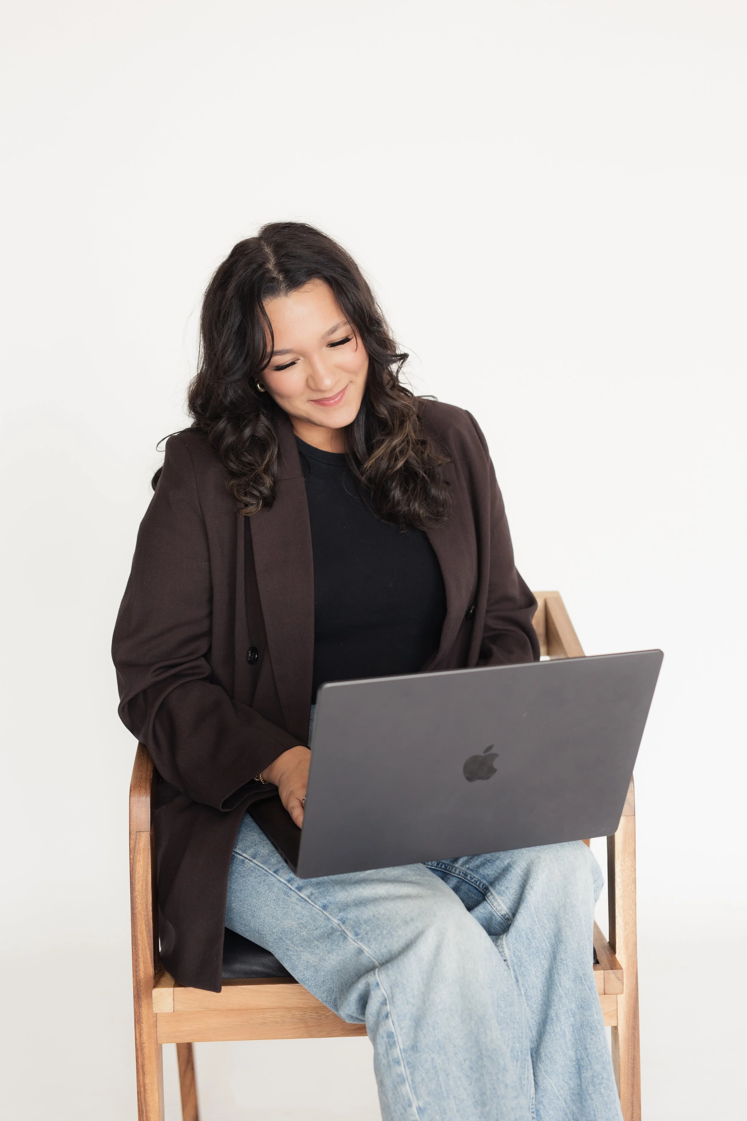 A woman with dark, wavy hair wearing a black top and brown blazer, sitting on a wooden chair using a silver MacBook laptop, against a plain white background. Heirloom Media Co branding photoshoot