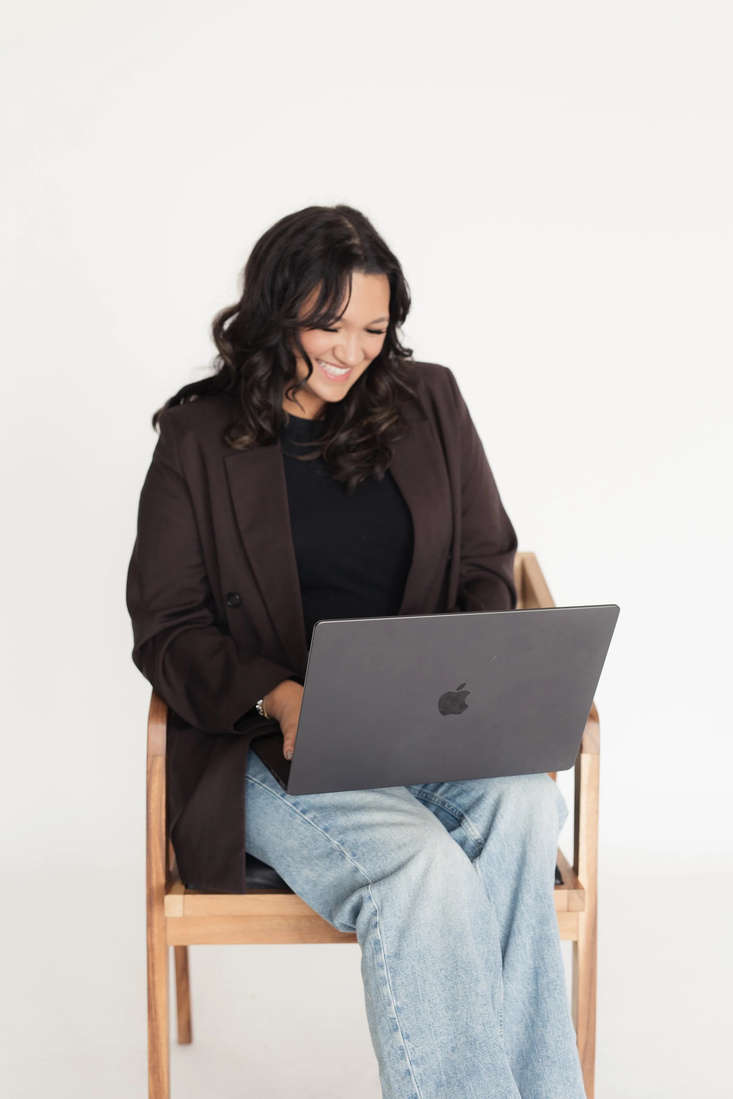 A woman sitting on a wooden chair, smiling and looking at her laptop, wearing a dark blazer and light blue jeans, against a plain white wall background. Heirloom Media Co branding photoshoot headshots
