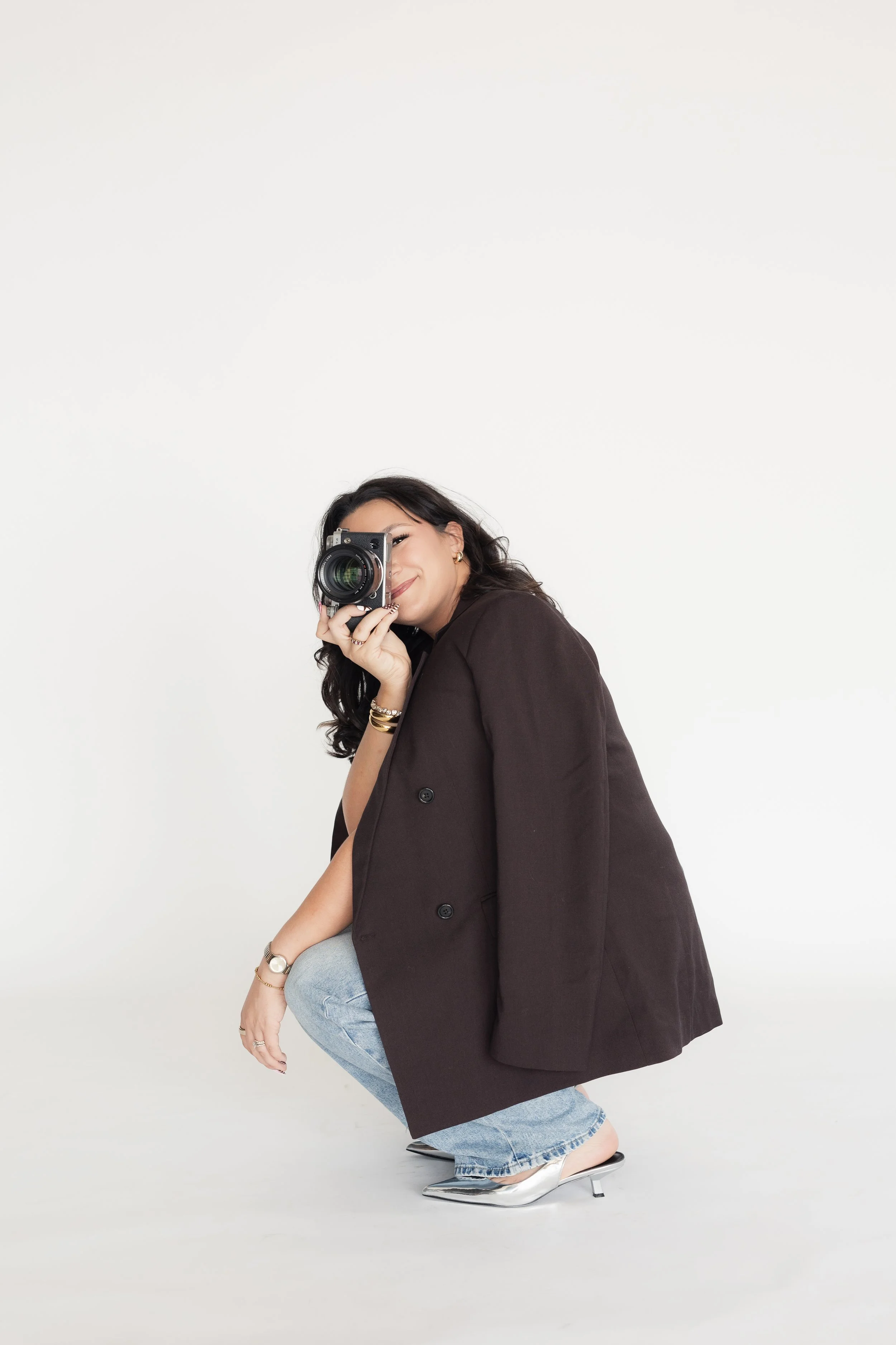 Woman crouching in a silver high heel shoe, wearing a dark blazer, light blue jeans, and jewelry, holding a camera up to her face against a plain white background. Heirloom Media Co branding photography