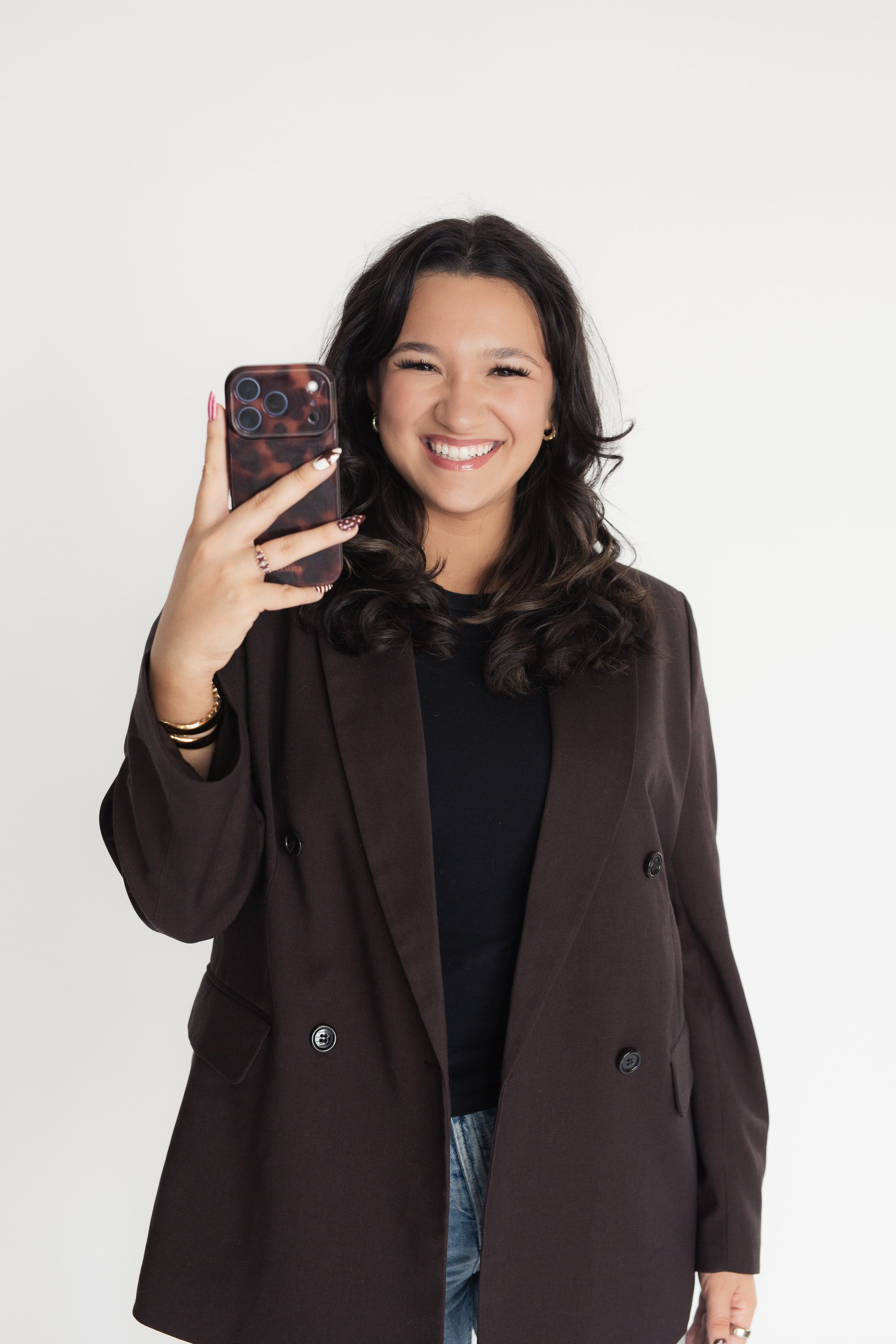A woman with dark wavy hair smiling, taking a photo with her smartphone, standing against a plain white background, wearing a dark blazer and jeans. Heirloom Media Co branding photoshoot and content creation