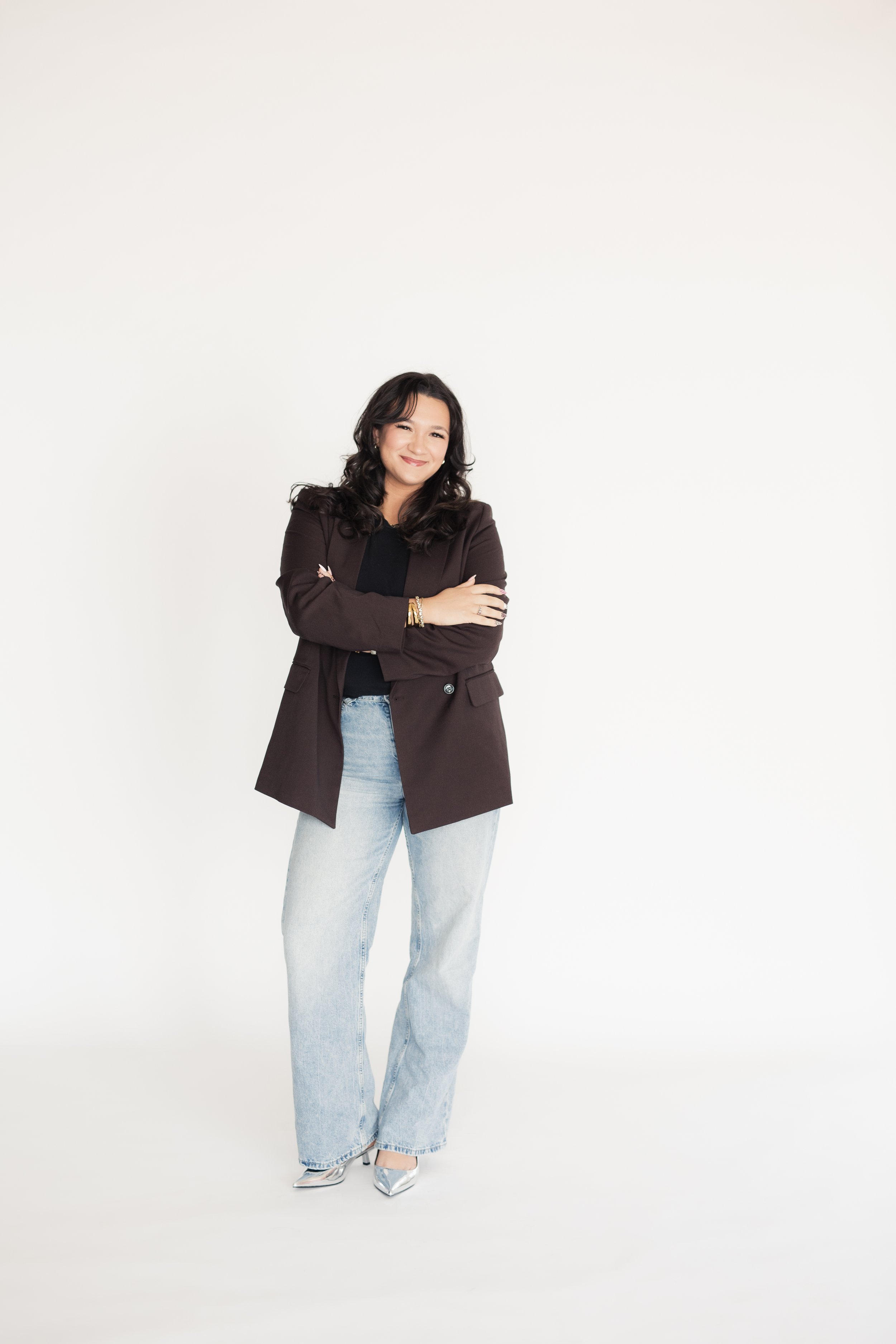 A woman with dark, wavy hair smiling, dressed in a black top, brown blazer, light blue jeans, and silver pointed shoes, standing with arms crossed against a plain white background. - Heirloom Media Co branding photoshoot