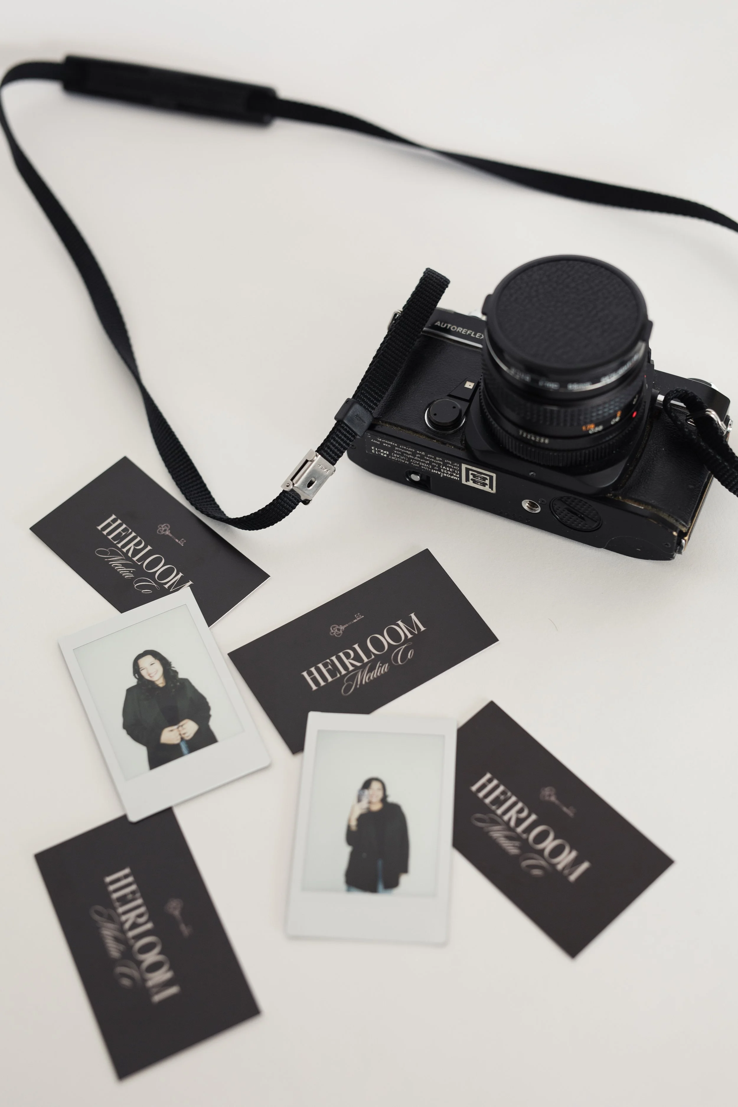 A black camera with a strap, several business cards from HEIRLOOM Media Co., and polaroids on a white surface. Heirloom Media Co branding photoshoot