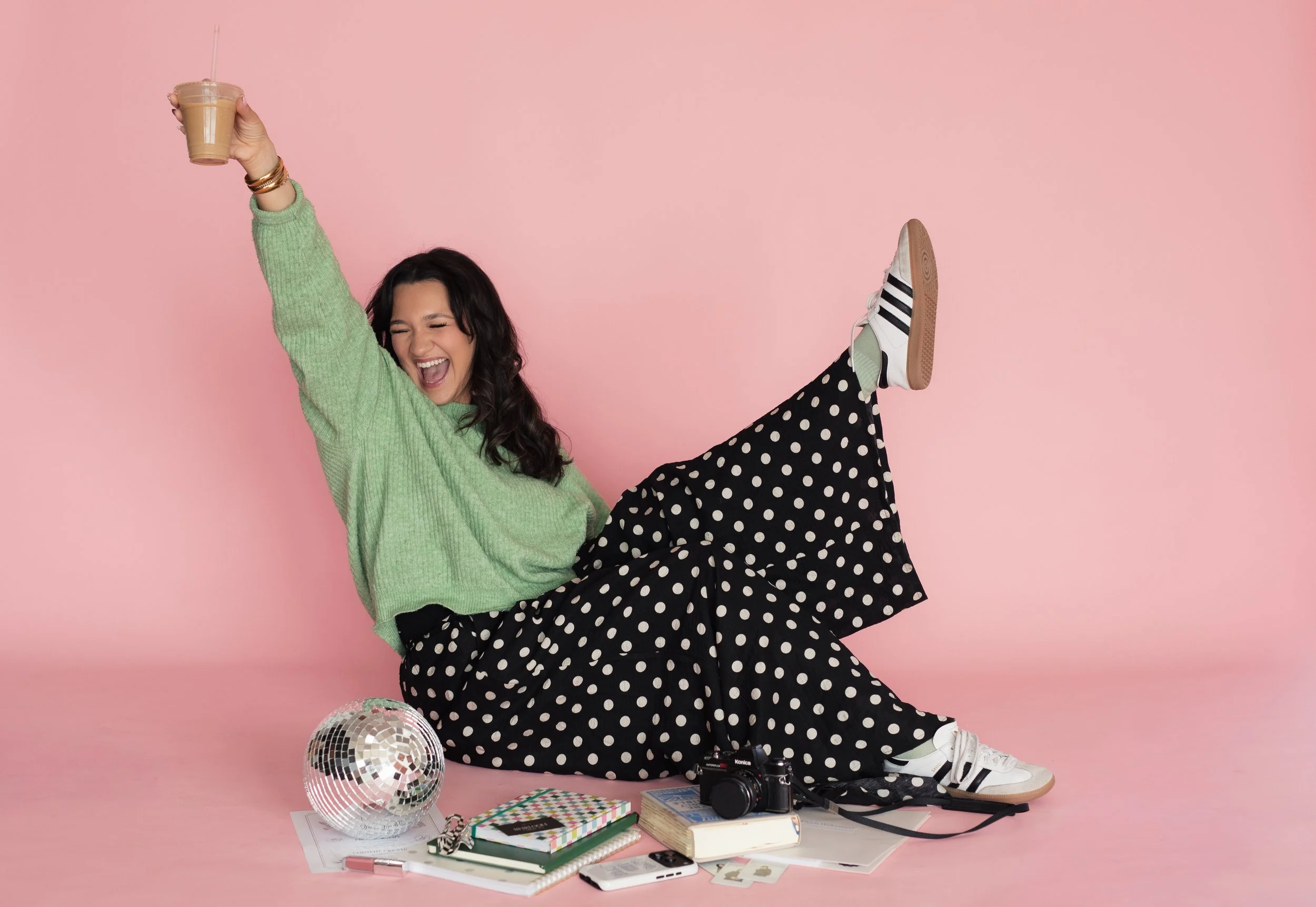 A young woman with long dark hair and a bright smile, wearing a green sweater and polka dot pants, is sitting on the floor against a pink background. She is holding a large iced coffee in one hand, raised excitedly, and has various items scattered around her including books, a camera, a disco ball, a smartphone, and some papers.