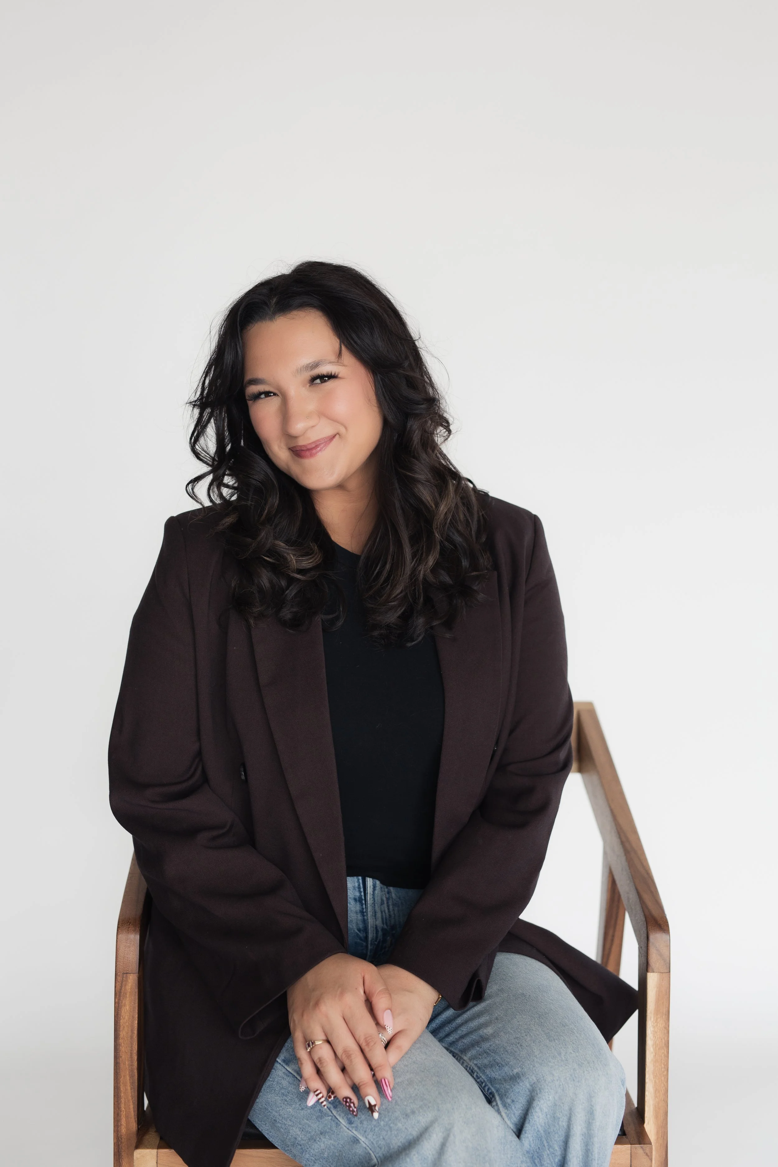 A woman with dark, wavy hair sitting on a wooden chair, smiling, wearing a black top, brown blazer, and light blue jeans, against a plain white background - Sarah, the founder of Heirloom Media Co's branding headshots