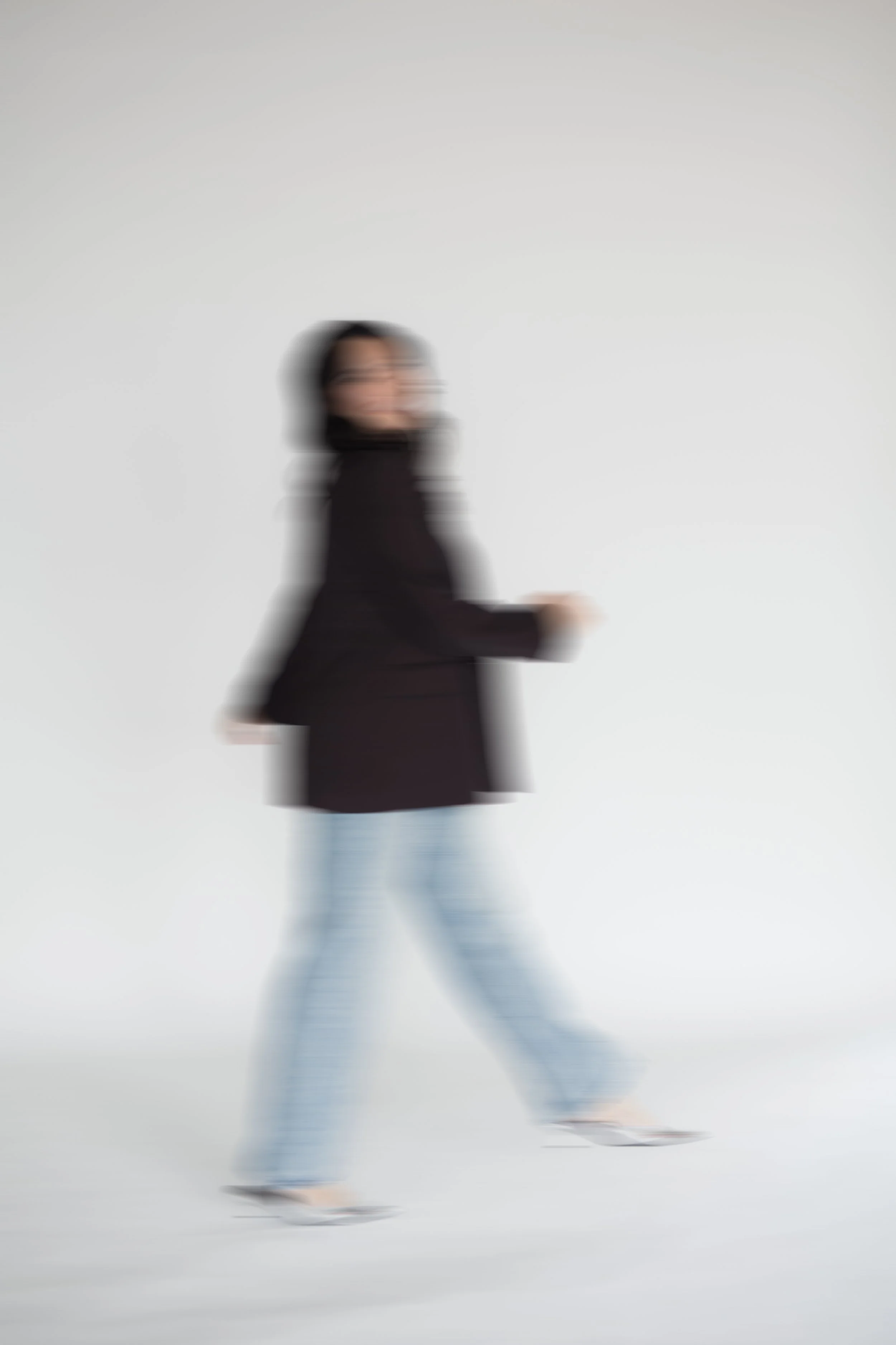 A woman with dark hair, wearing light jeans, silver shoes, and a dark brand blazer, is mid-motion and appears blurry against a plain light background. Heirloom Media Co branding photos