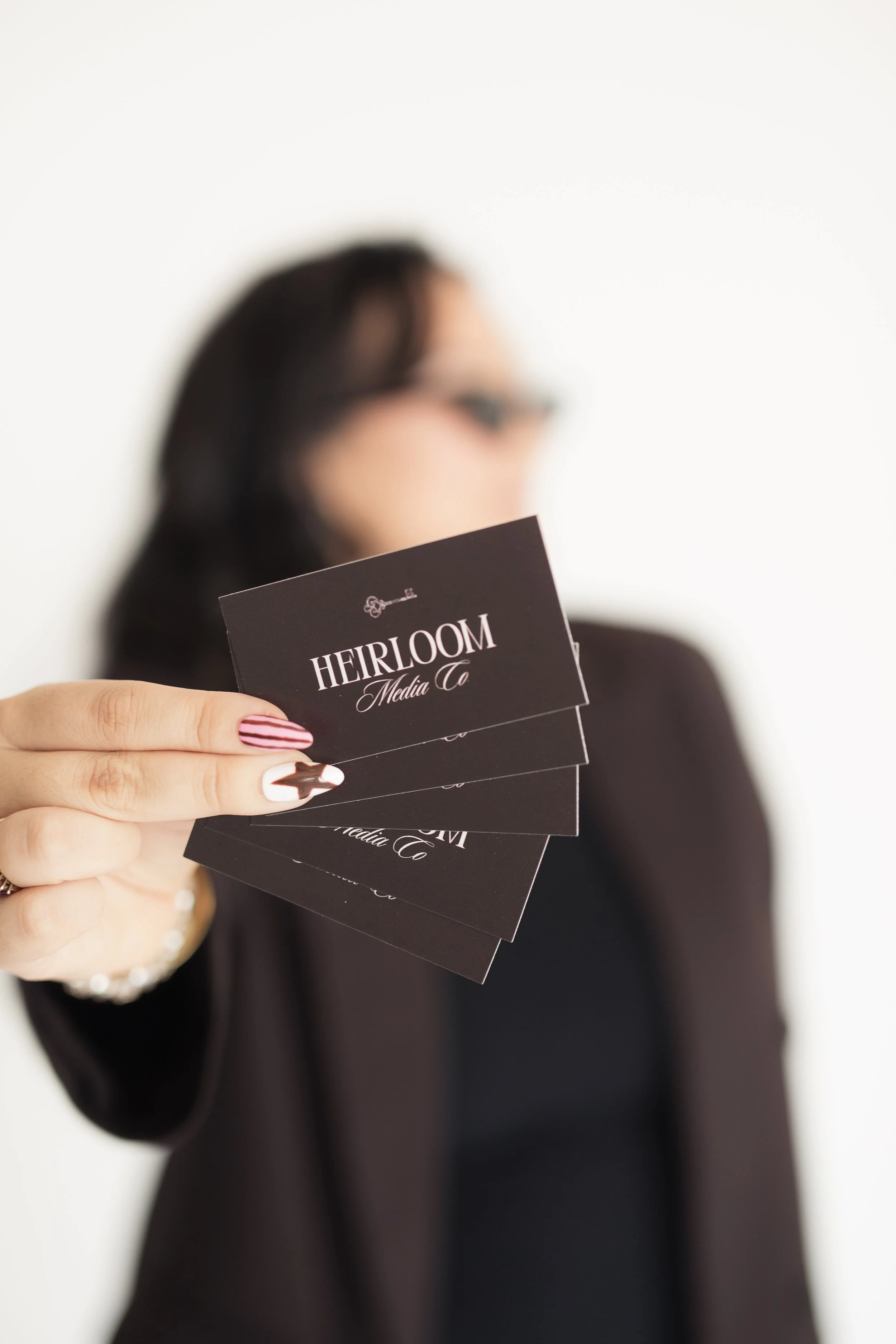 A woman with dark hair and glasses holding business cards that say "HEIRLOOM Media Co." in front of a plain white background. Heirloom Media Co branding photoshoot