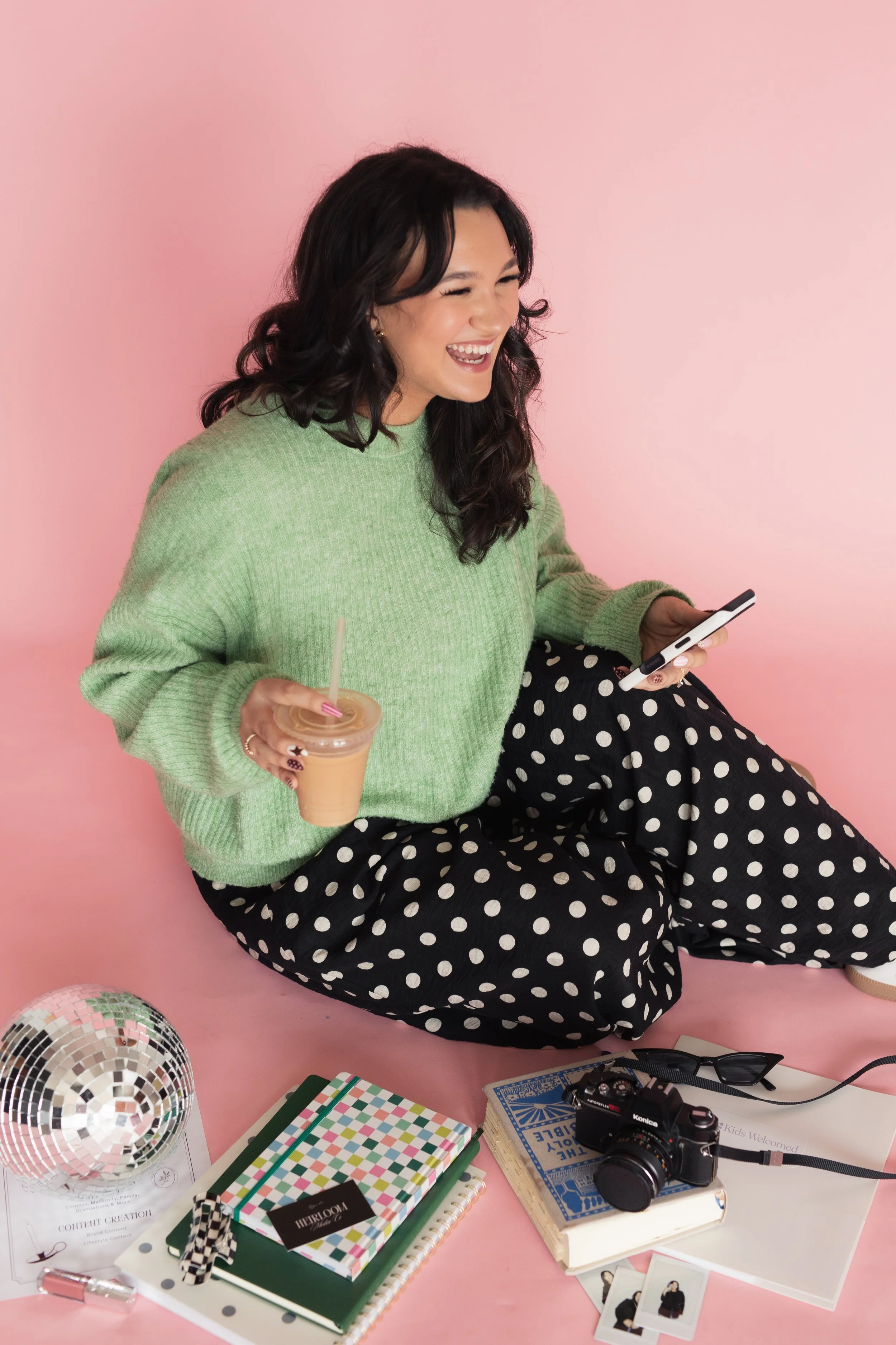 A young woman with dark, curly hair, wearing a green sweater. She is smiling and looking at her phone, holding a drink with a straw. In front of her are various items, including a color swatch book, notebooks, a camera, polaroid photos, and a small disco ball.