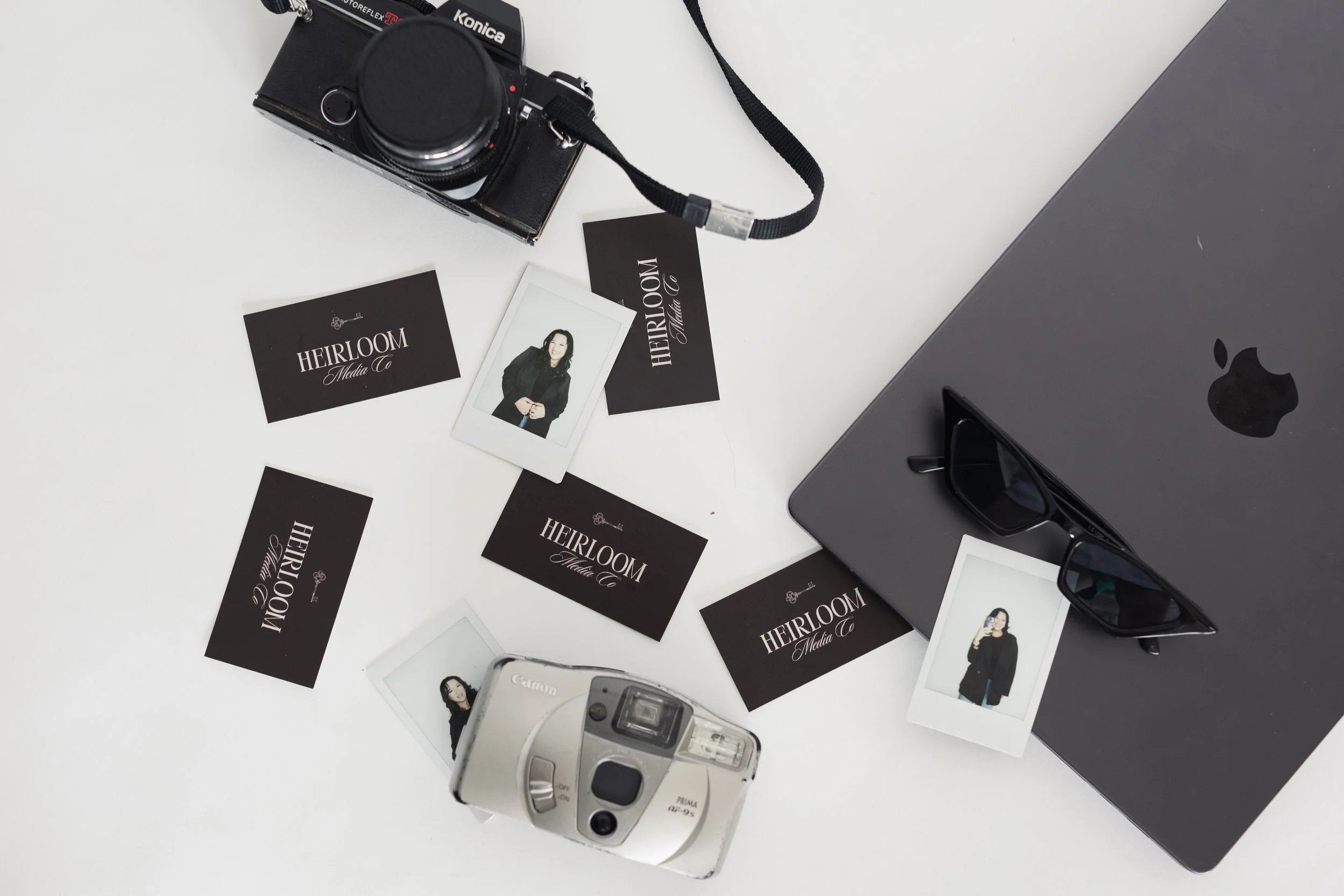 A camera, a computer, business cards for Heirloom Media Co, a smartphone, and sunglasses on a white background. Heirloom Media Co branding photoshoot.