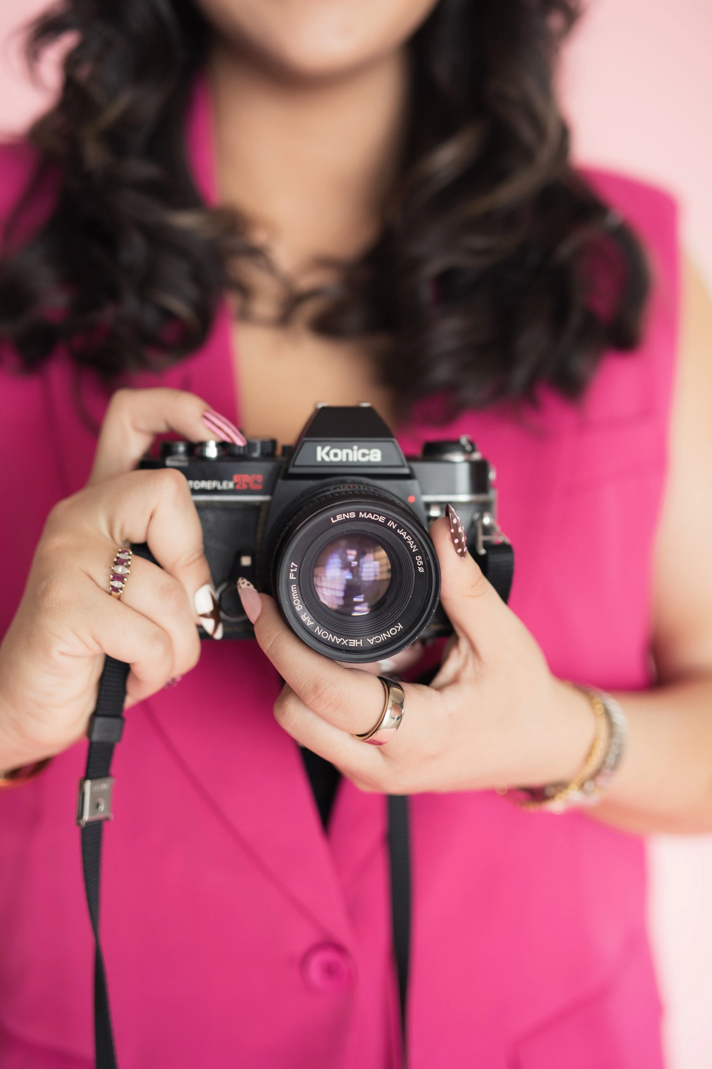 A woman in a pink blazer vest holding a vintage Konica film camera, with curled dark hair and jewelry on her fingers and wrist. Heirloom Media Co branding photoshoot