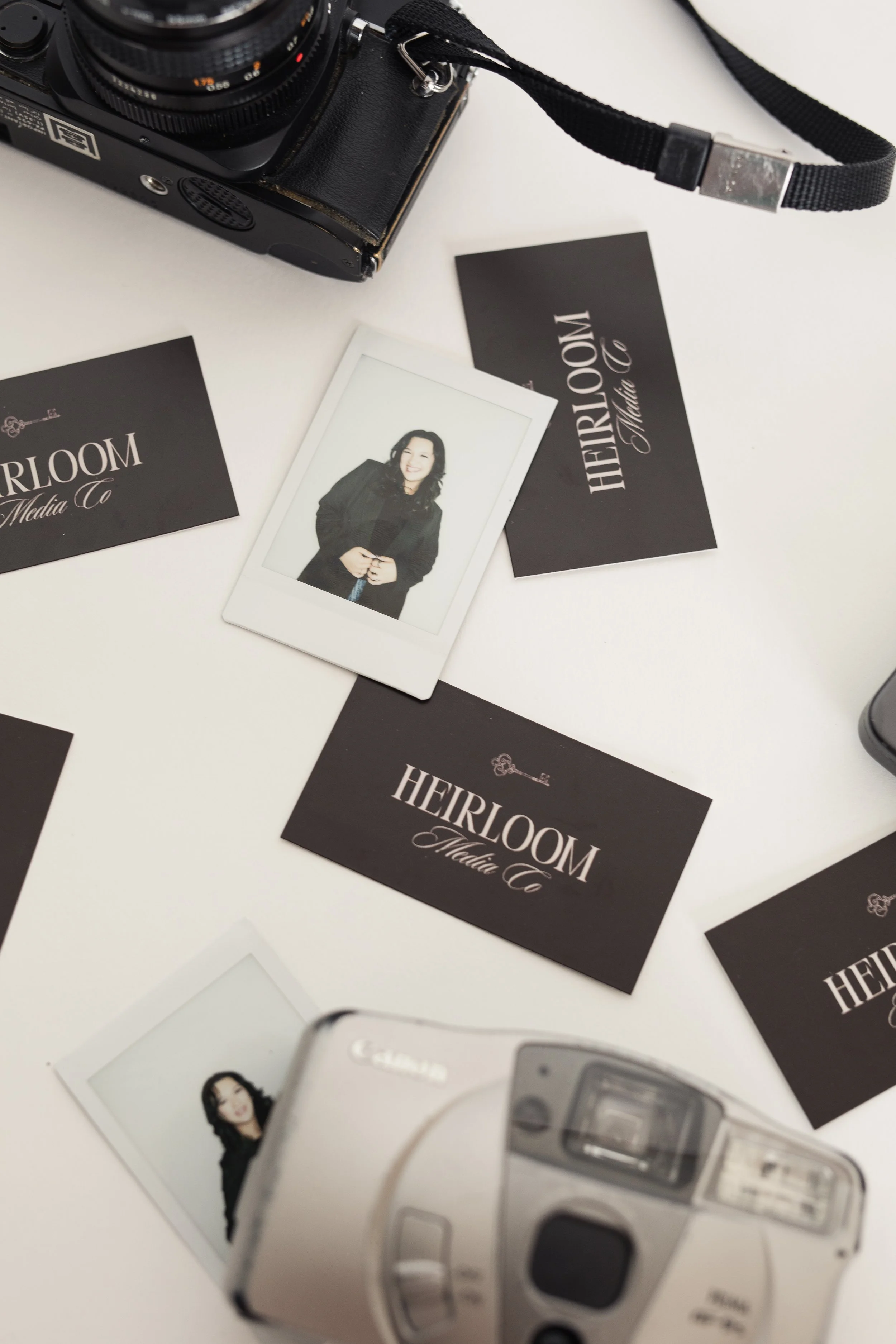 Two film cameras, business cards reading 'HEIRLOOM Media Co', and a Polaroid of a woman on a white surface.