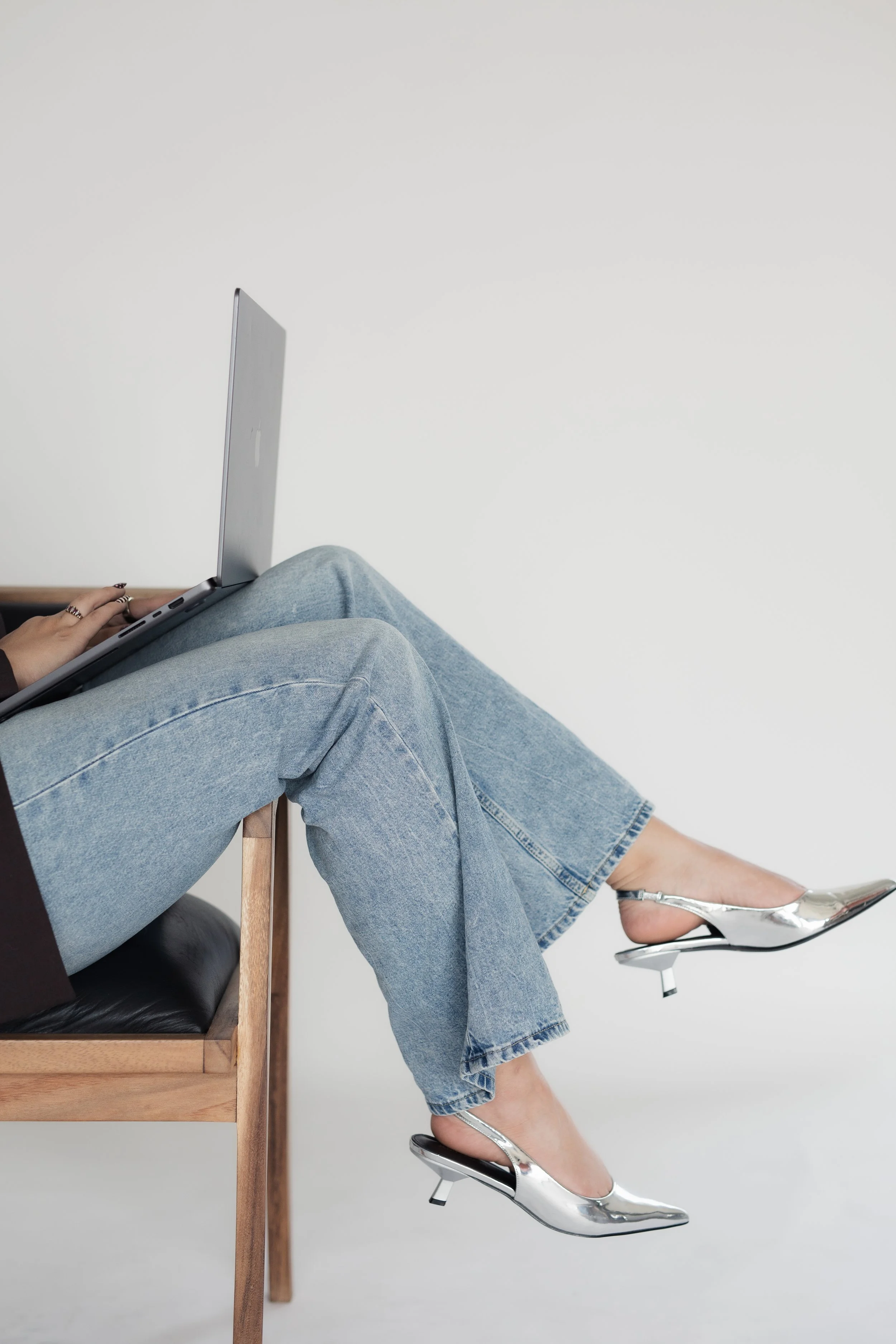 A person sitting on a black leather and wood chair with their legs crossed, wearing light blue jeans, a black top, and metallic high-heeled shoes, with a computer on their lap, against a plain white background.