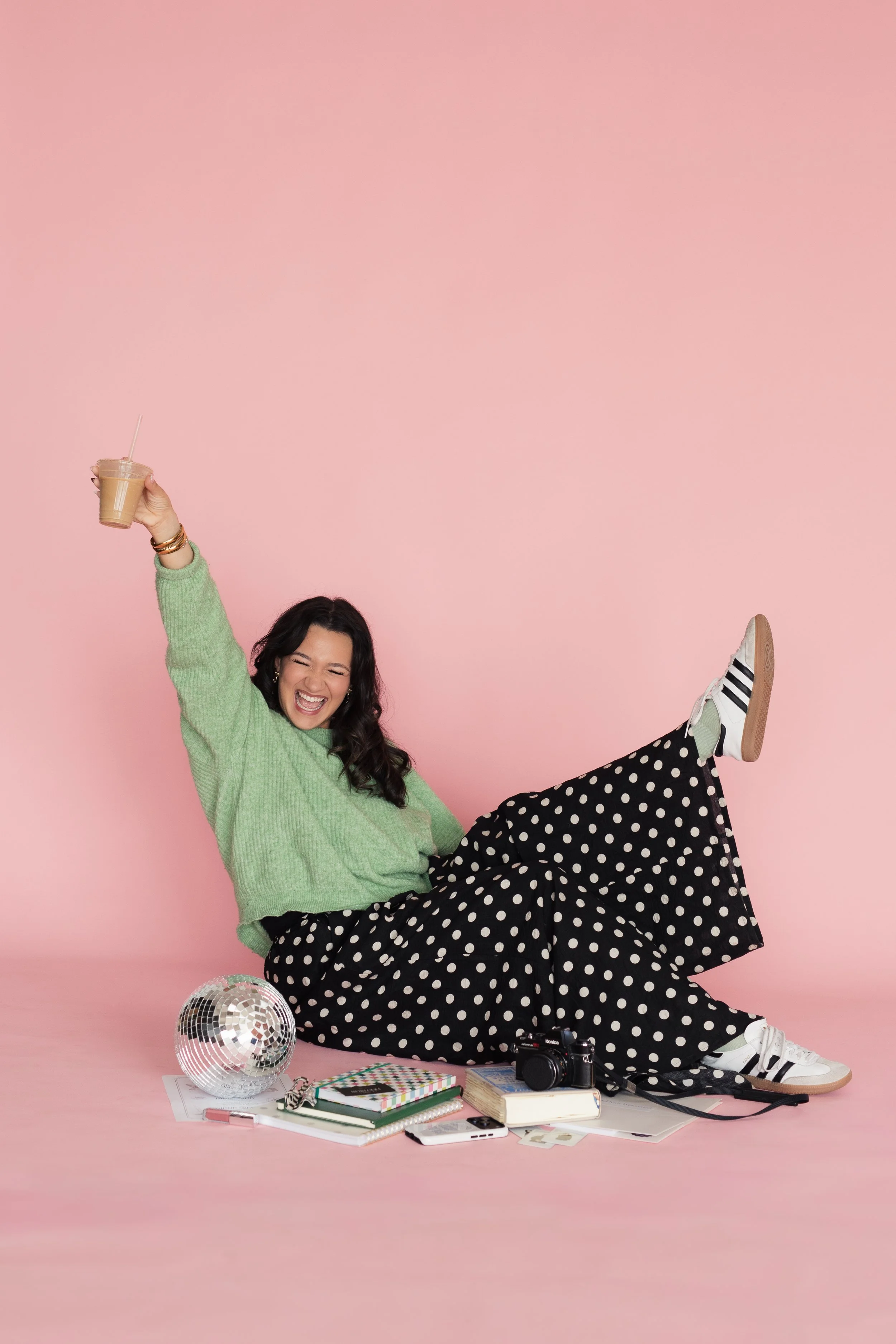 A young woman with dark hair, dressed in a green sweater and black polka dot pants, sitting on the floor against a pink background. She is smiling, raising one arm holding a coffee cup, with various items like notebooks, a camera and a disco ball.