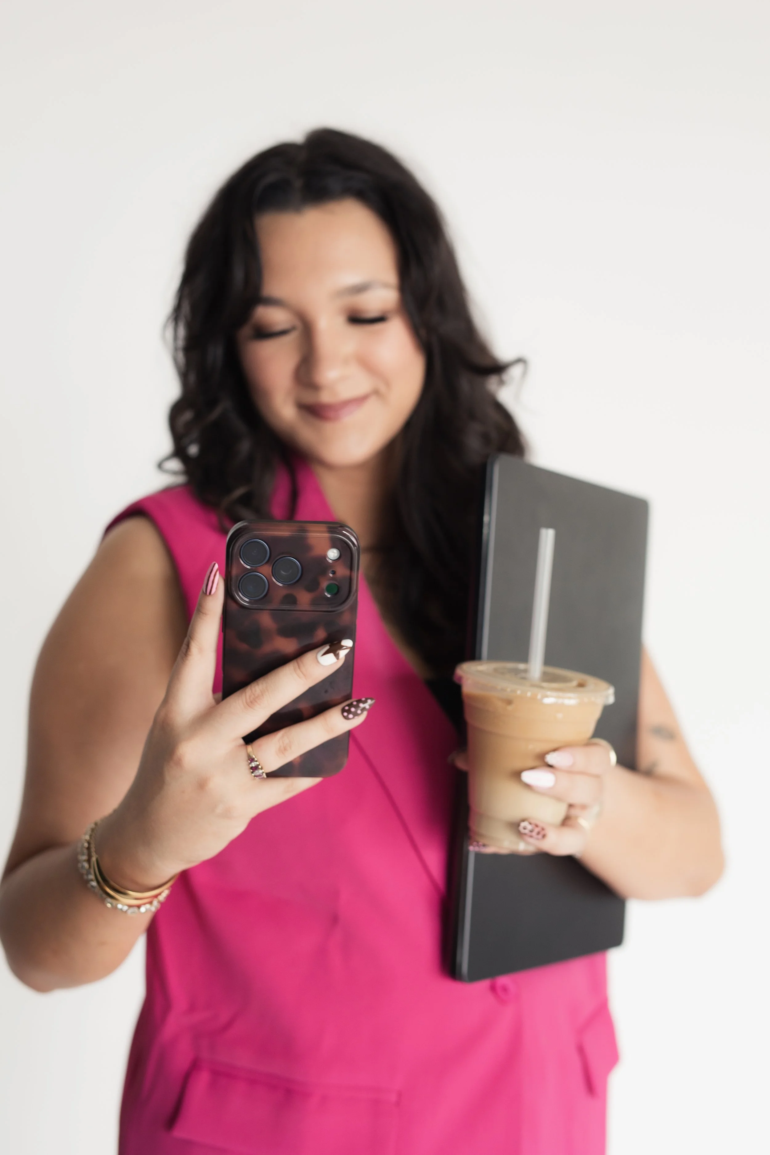 A woman with dark, curly hair wearing a pink top holds a smartphone in her right hand and a coffee drink in her left hand, with a laptop resting against her torso. Heirloom Media Co branding photoshoot. Content creation by Heirloom Media Co