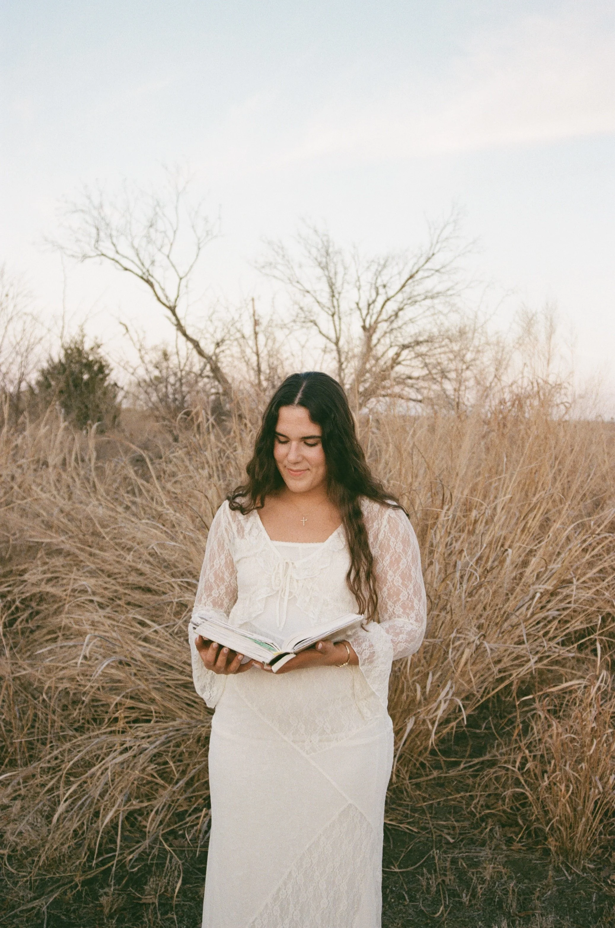 A young woman with long dark hair in a white lace dress, holding an open Bible, standing in a field of tall, dry grass with leafless trees and a cloudy sky in the background.