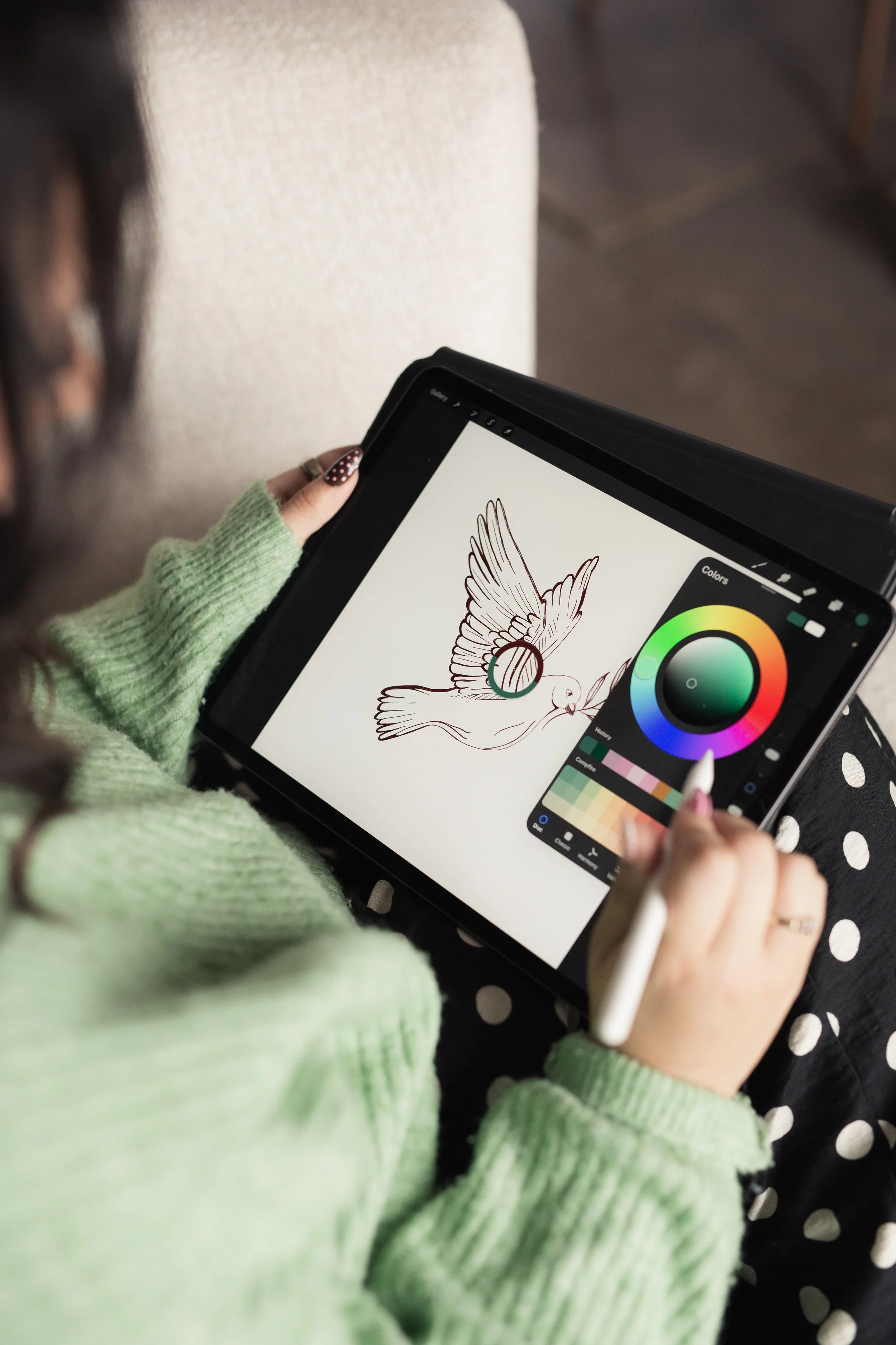 A person wearing a green sweater and black and white polka dot pants, sitting on a large cozy chair, using a stylus on a tablet to color a drawing of a bird, with color palette and tools visible on the screen.