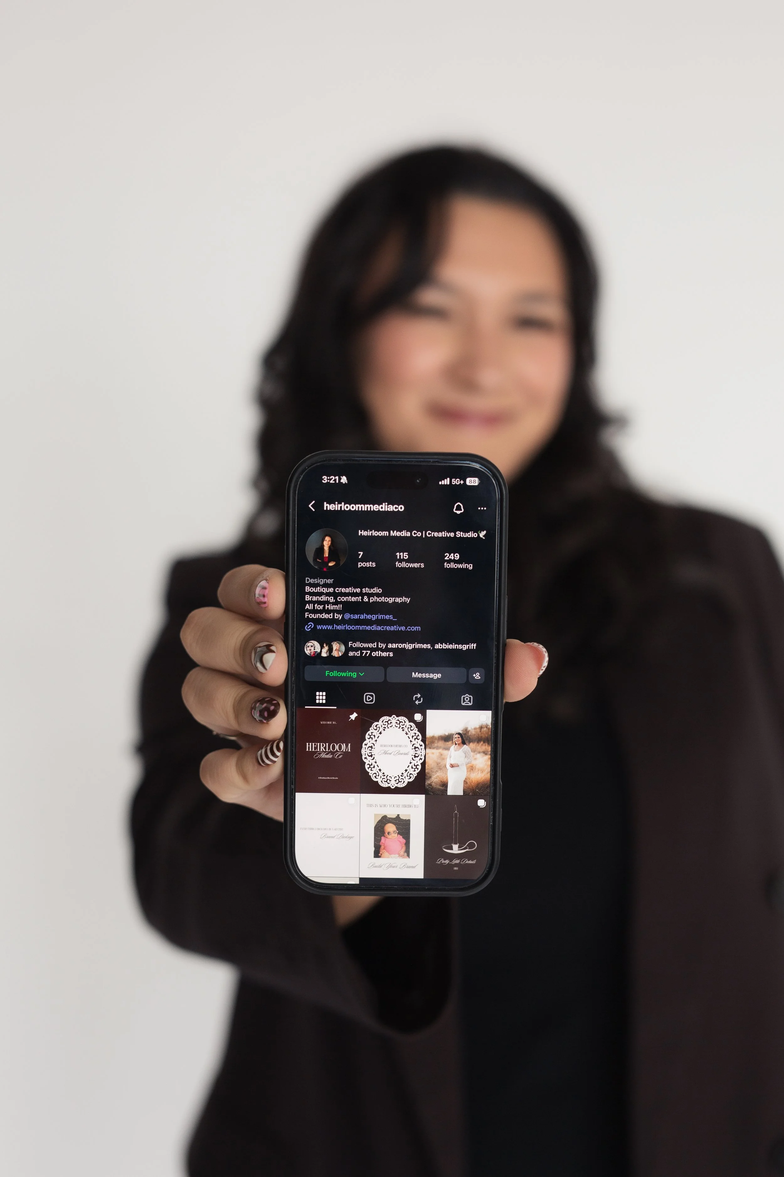 A woman with dark hair and nail art is holding a smartphone displaying her social media profile, Heirloom Media Co, with various images and posts visible. Heirloom Media Co branding photoshoot