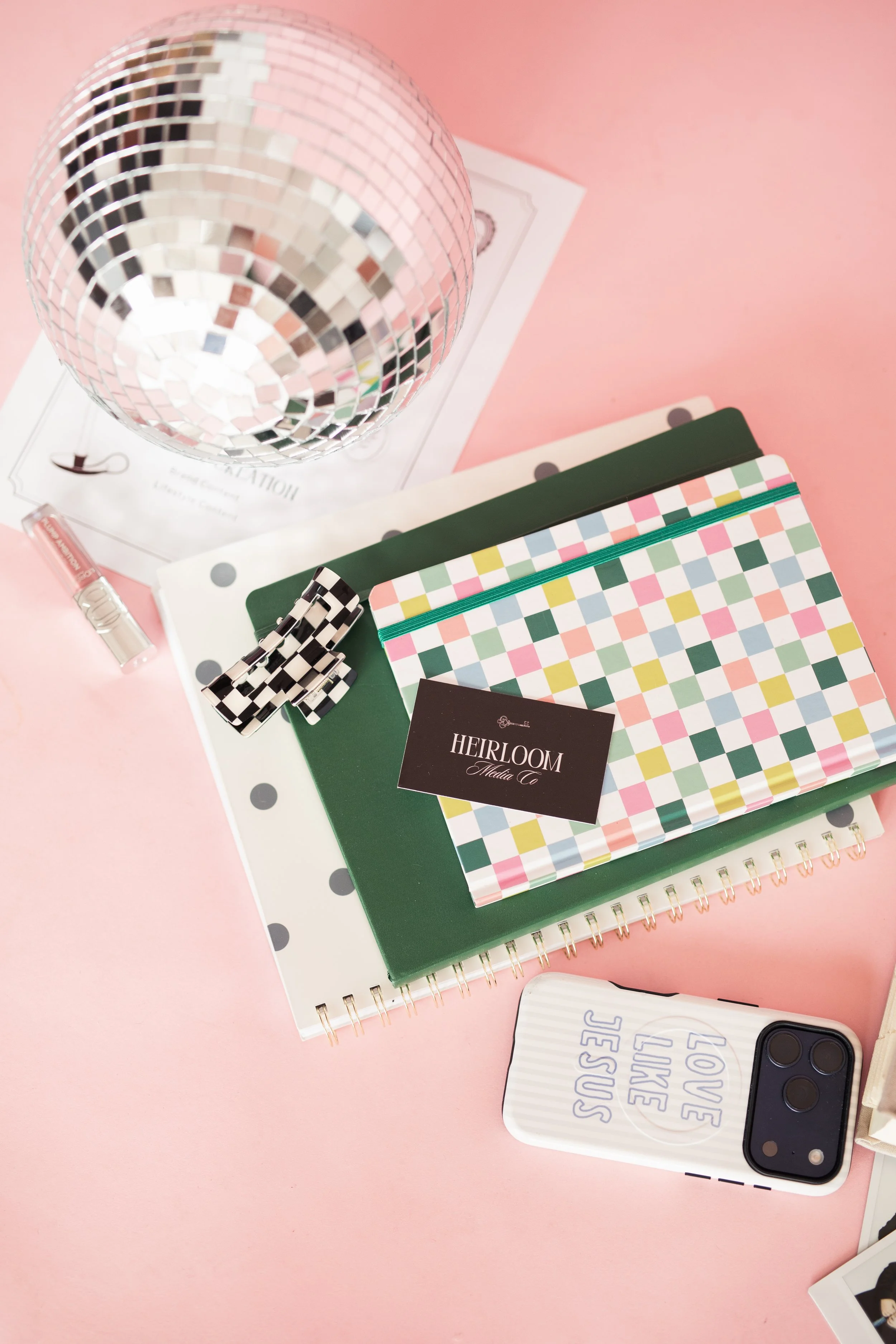 A flat lay of assorted stationery items on a pink background, including a disco ball, notebooks with colorful checkered covers, a white organizer with gray polka dots, a hair clip, a lipstick, a business card, and a smartphone with a striped case.