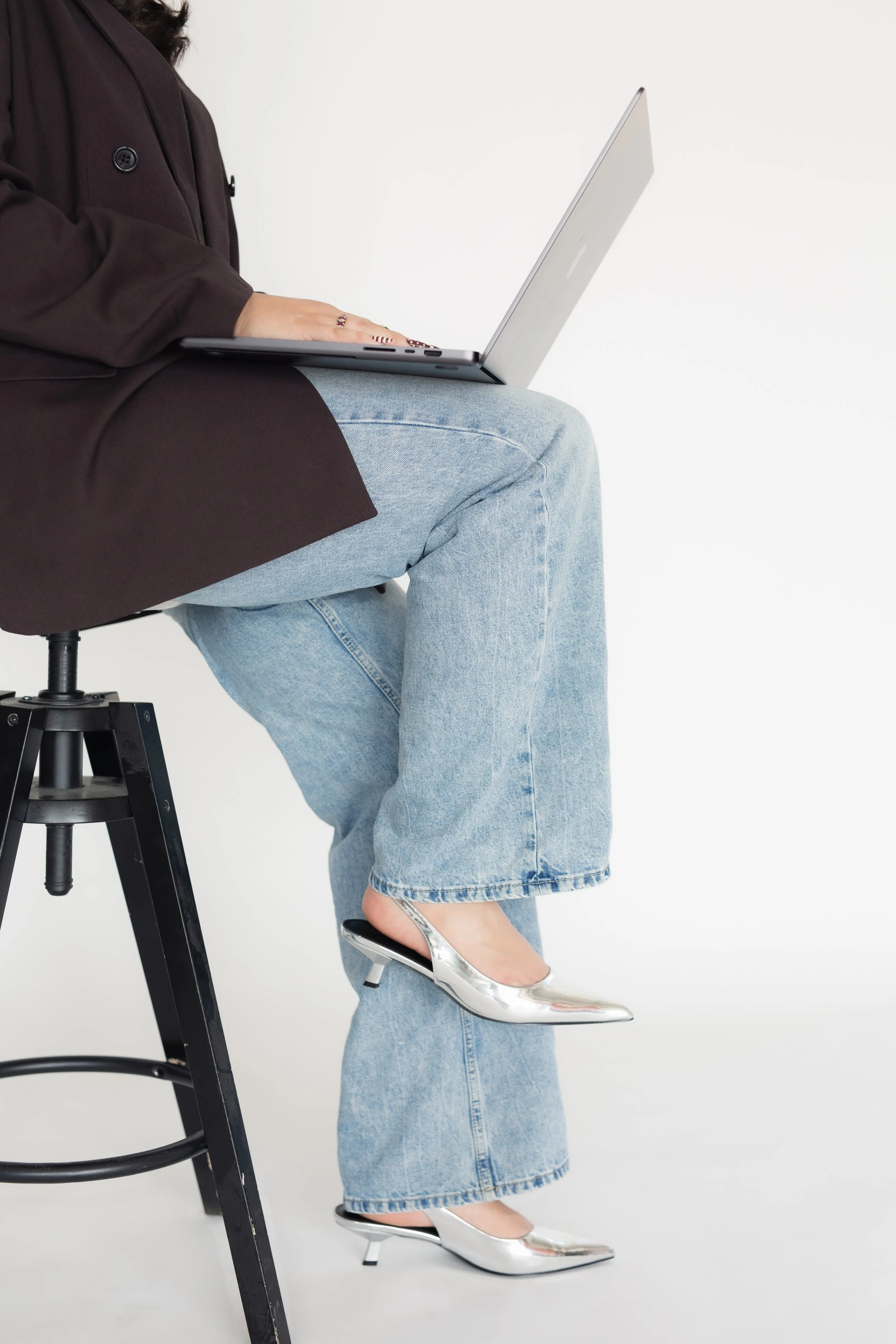 A person sitting on a stool with one leg crossed over the other, wearing high heels, blue jeans, and a dark coat, with a laptop on their lap against a plain white background.