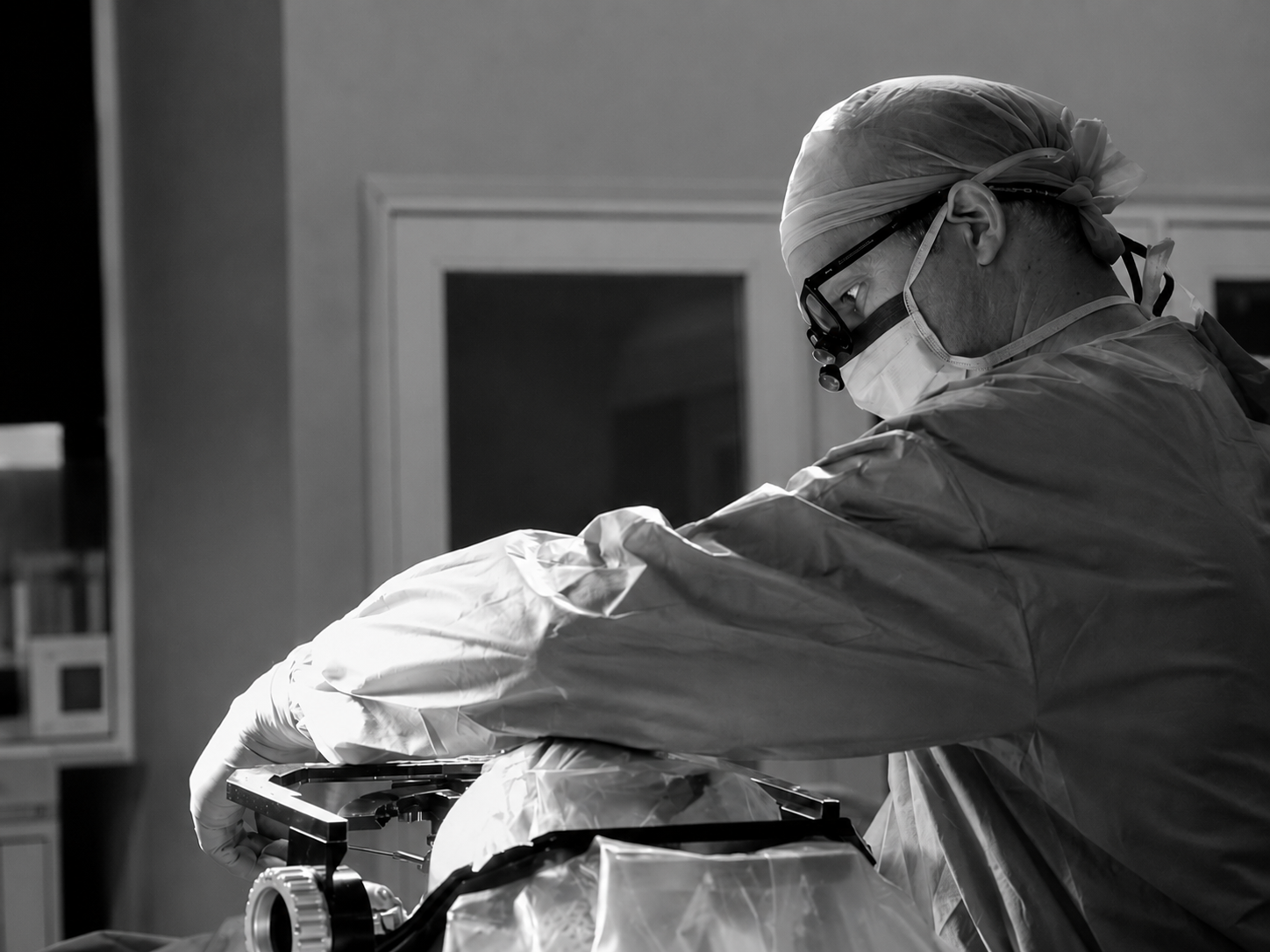A surgeon wearing a surgical cap, mask, and goggles operates on a patient in a hospital operating room.