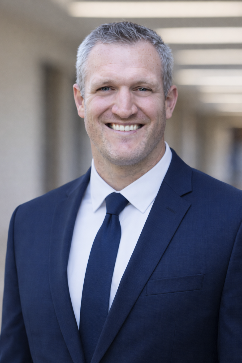 A smiling man in a navy suit, white shirt, and navy tie standing outdoors, with a blurred background of a building corridor.