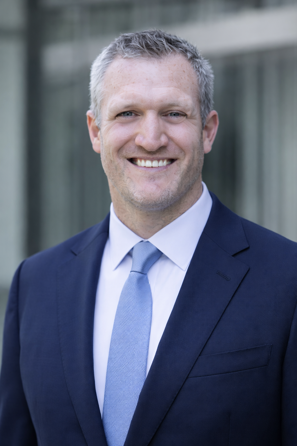 A man in a dark suit, white shirt, and light blue tie smiling outdoors with a blurred background.