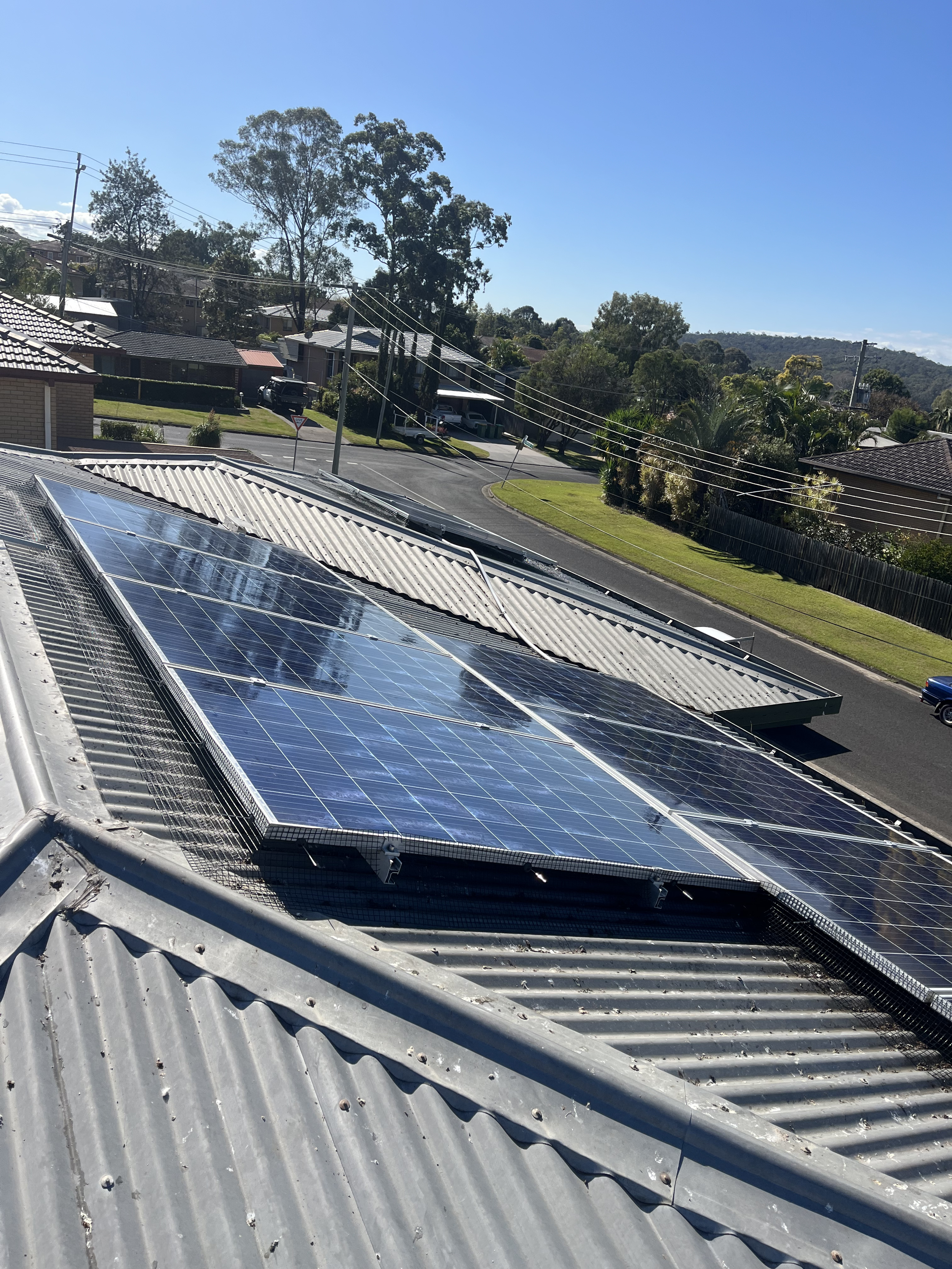 Solar panels installed on corrugated metal roof in a suburban neighborhood with trees and houses in the background.