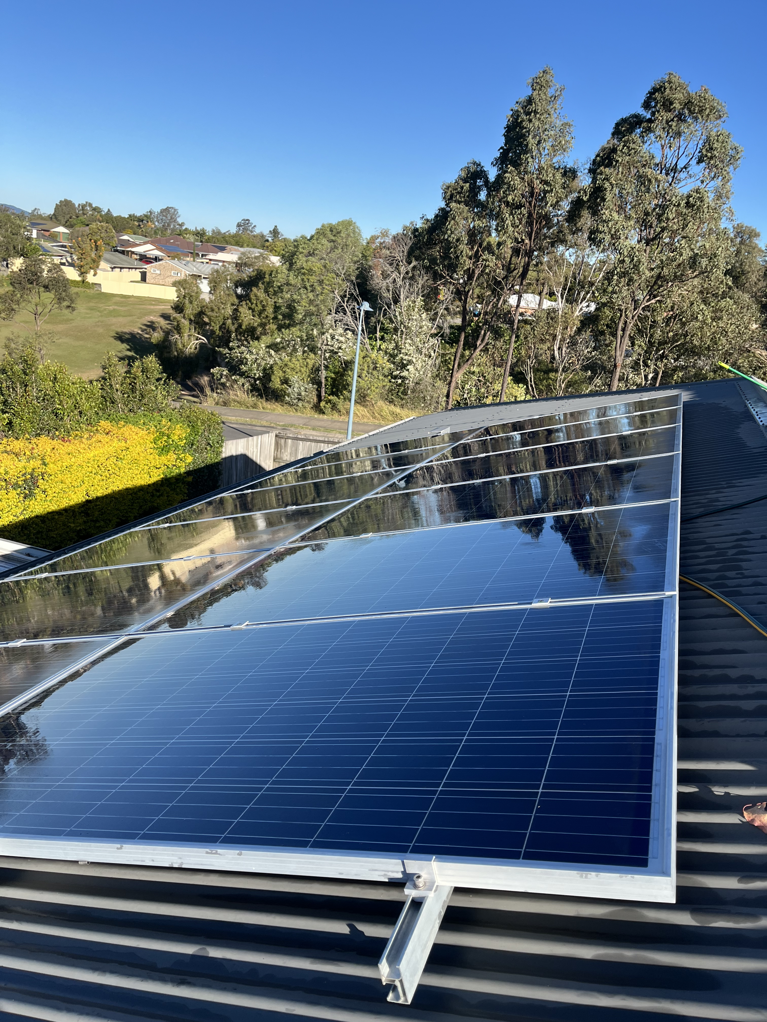 clean Solar panels after being washed on a metal roof, with a background of trees, houses, and a clear blue sky.