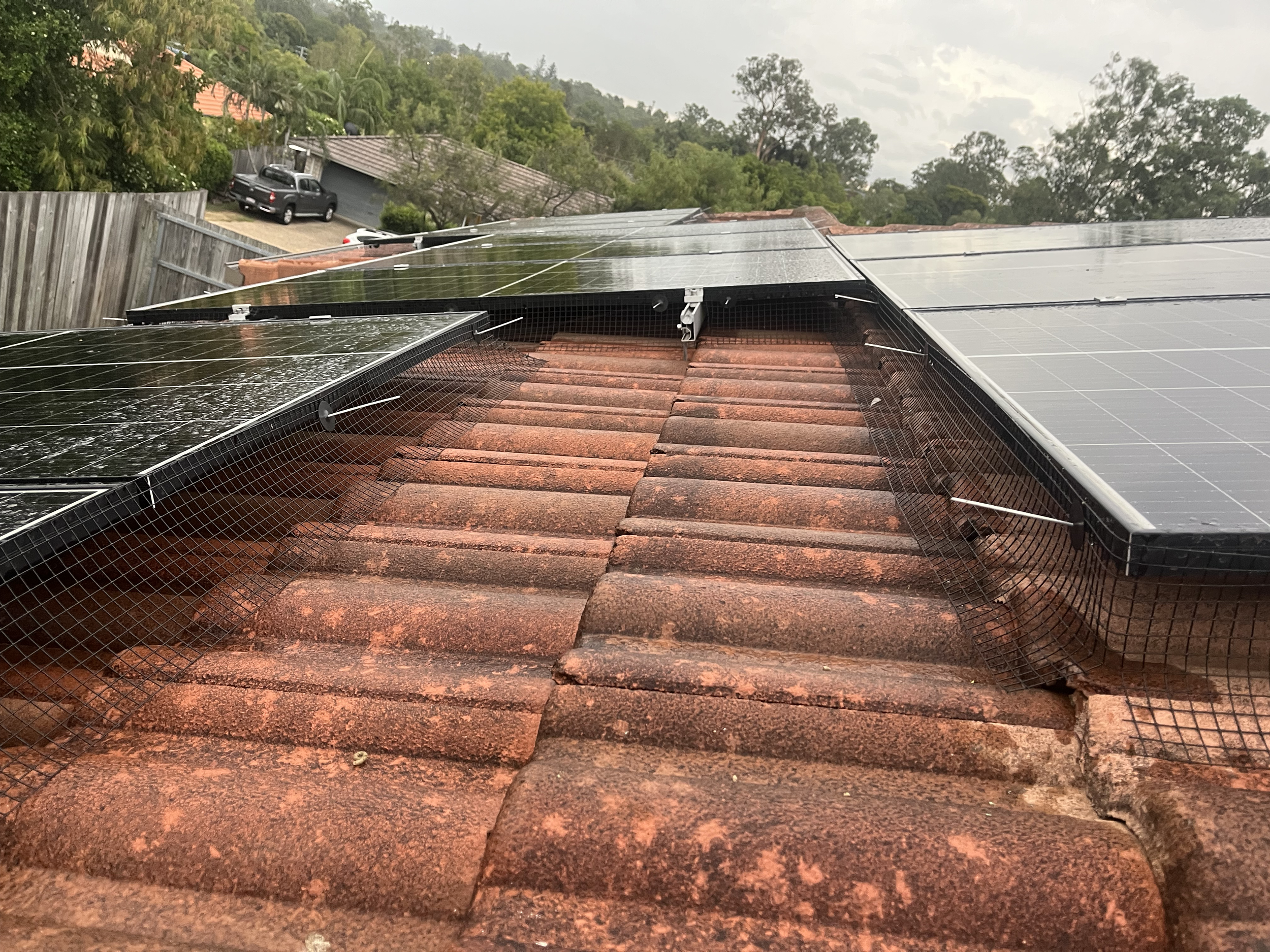 Solar panels with pigeon mesh installed on a tiled roof, with a background of trees, parked cars, and neighboring houses under a cloudy sky.