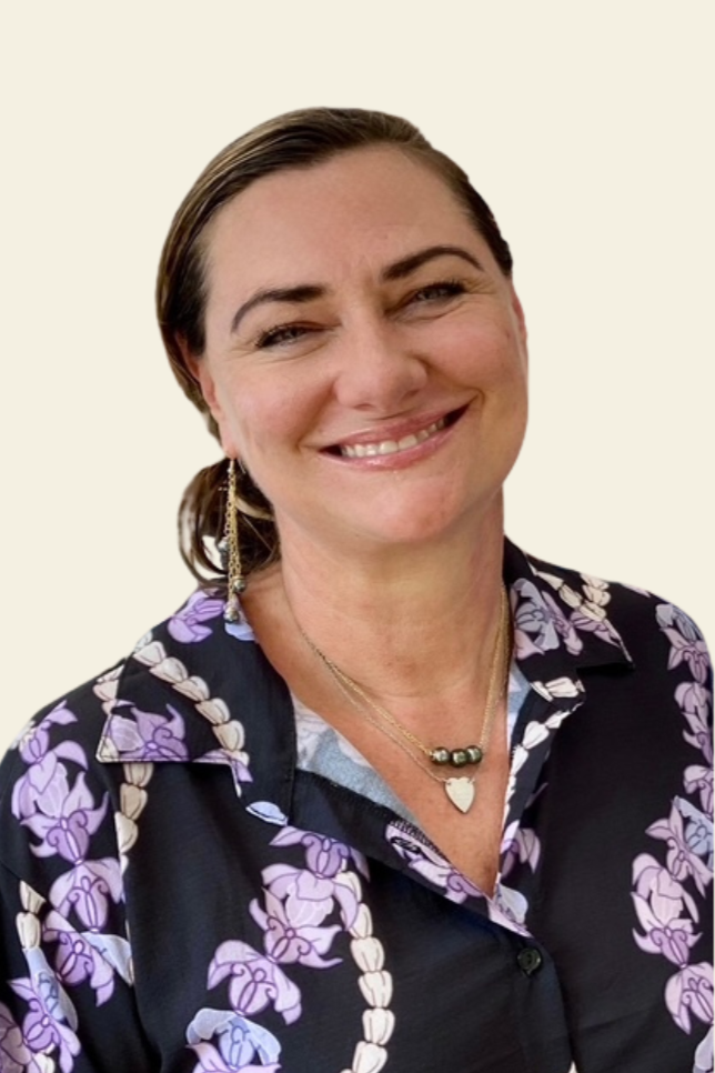 A woman smiling, wearing a patterned black shirt with purple and cream floral designs, gold heart necklace, and matching earrings, with dark brown hair pulled back, against a plain light-colored background.