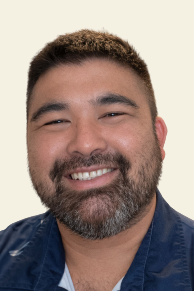Close-up of a smiling man with a beard and short dark hair, wearing a dark blue shirt, against a plain light background.