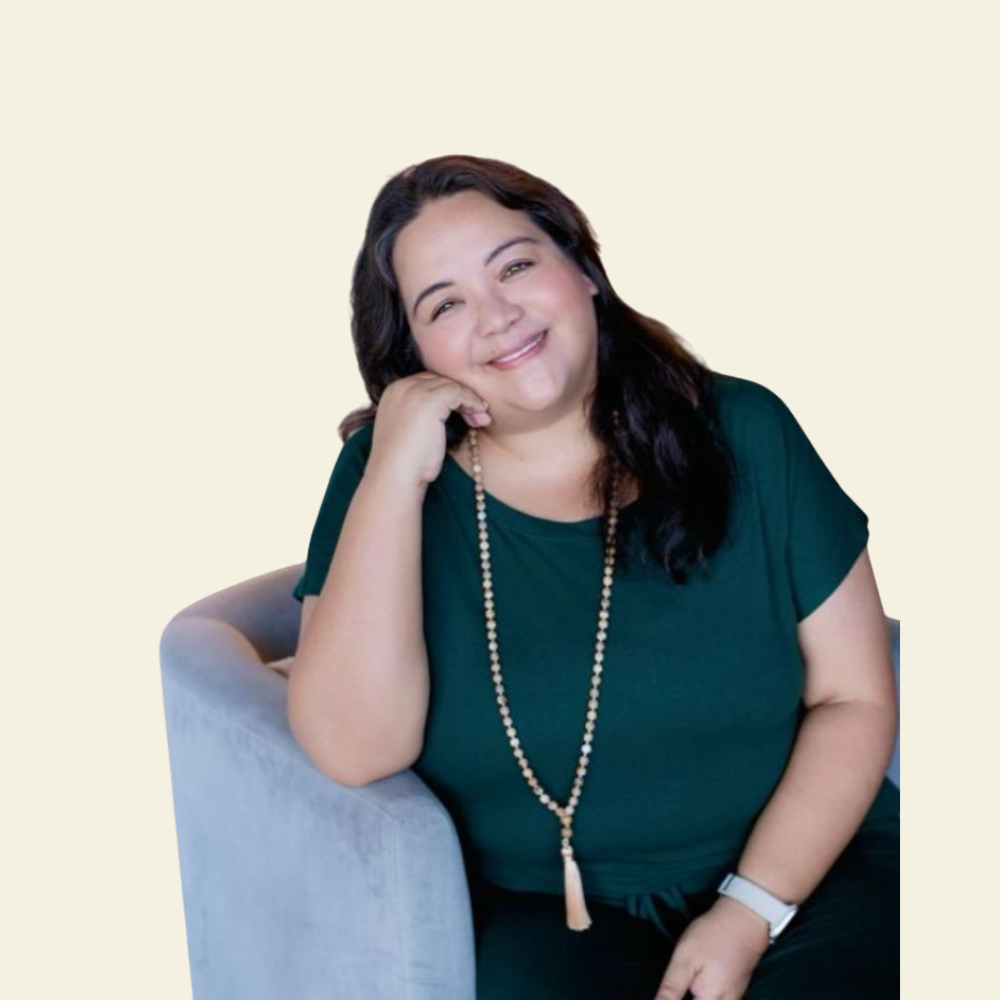 A woman with dark hair, wearing a teal shirt and a long beaded necklace, sitting on a light-colored couch with a cheerful expression, resting her head on her hand.