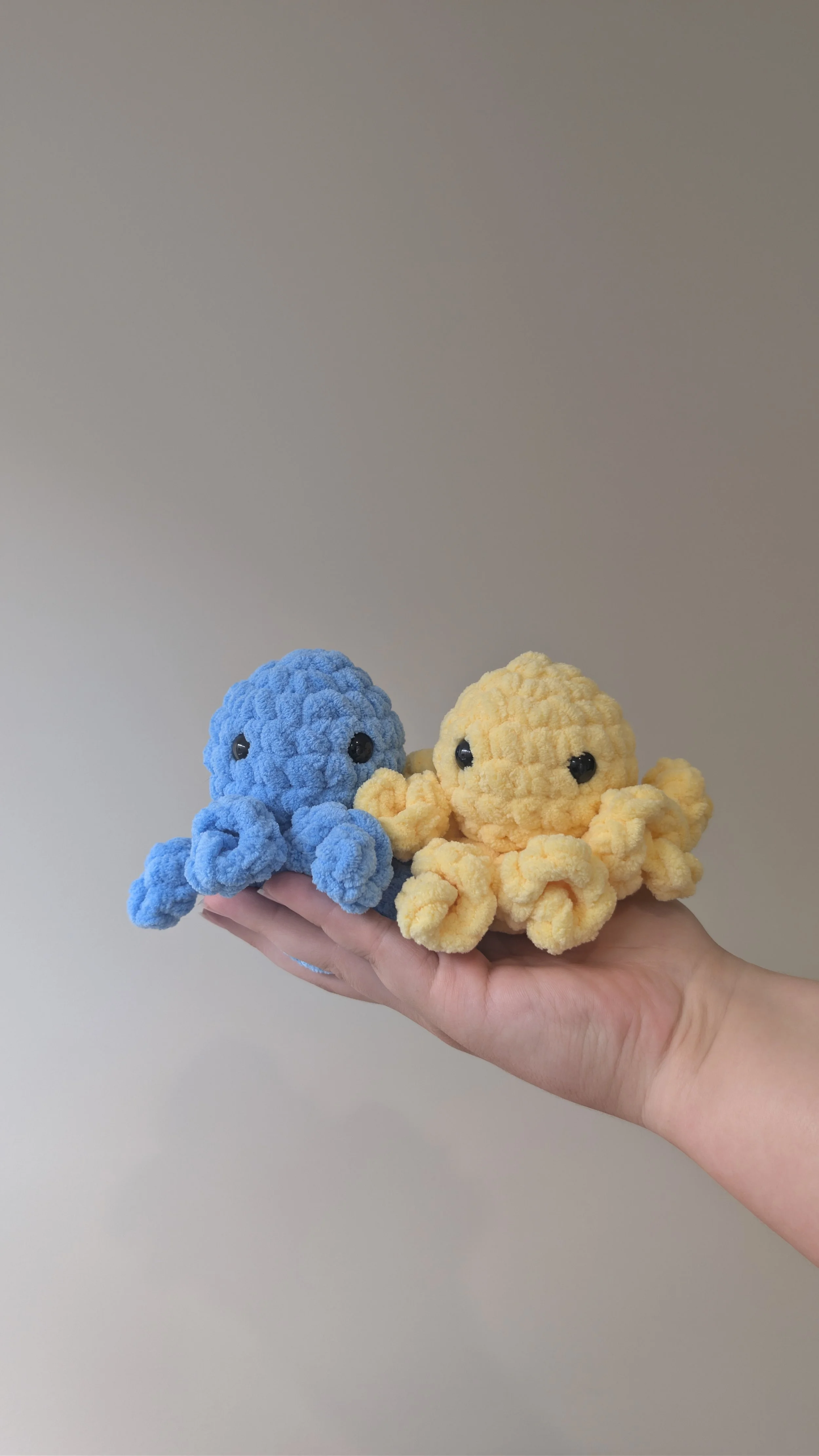 Hand holding two plush octopus toys, one blue and one yellow, with a plain beige background.