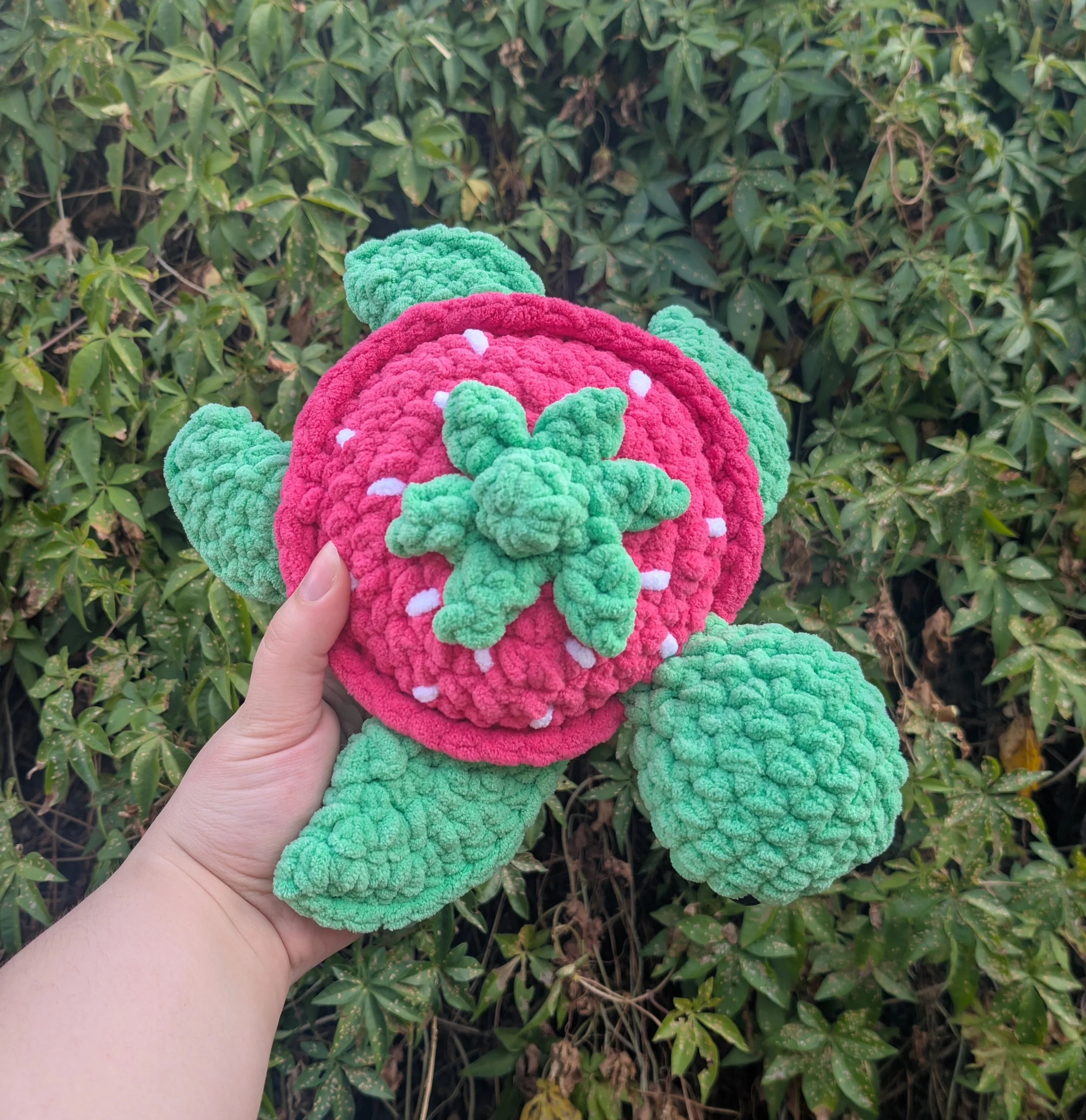 Person holding a plush toy designed to look like a pink raspberry plant with green leaves, set against a background of green leafy bushes.