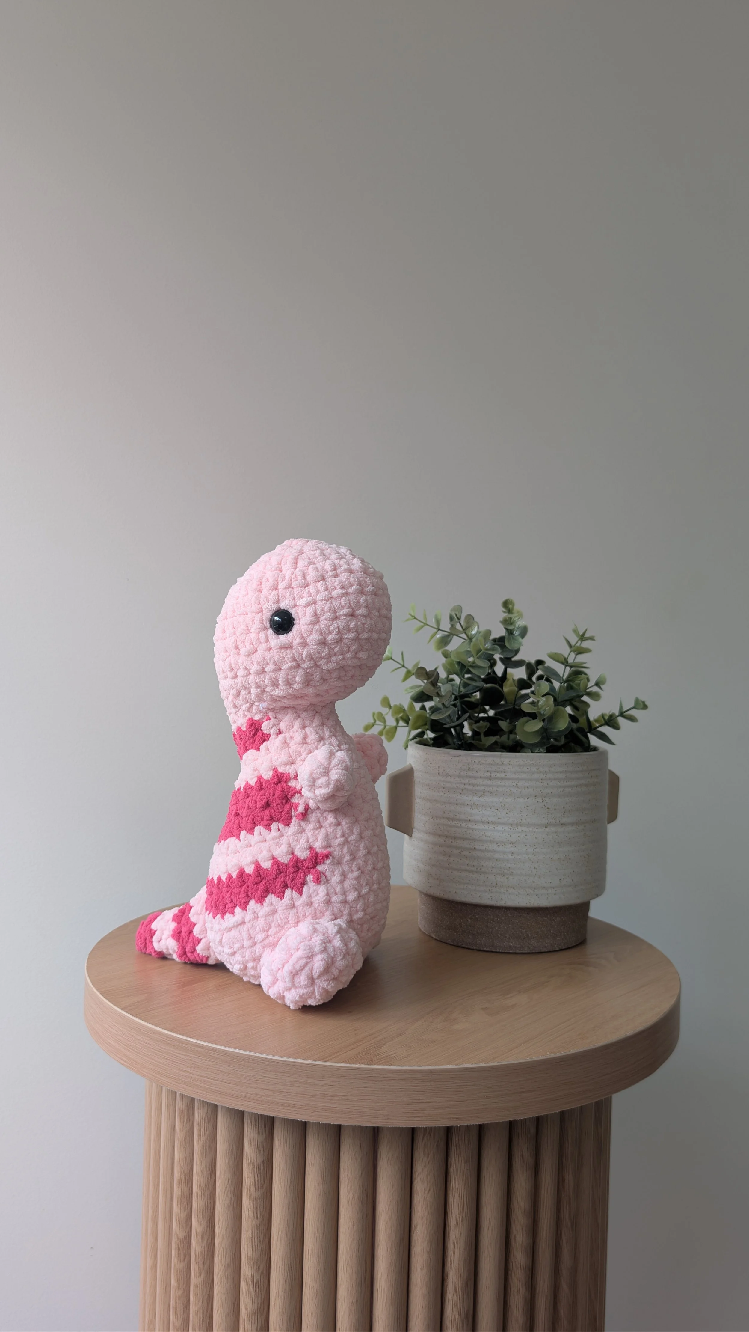 A crocheted pink plush toy dinosaur with darker pink stripes sits next to a potted green plant on a wooden side table.