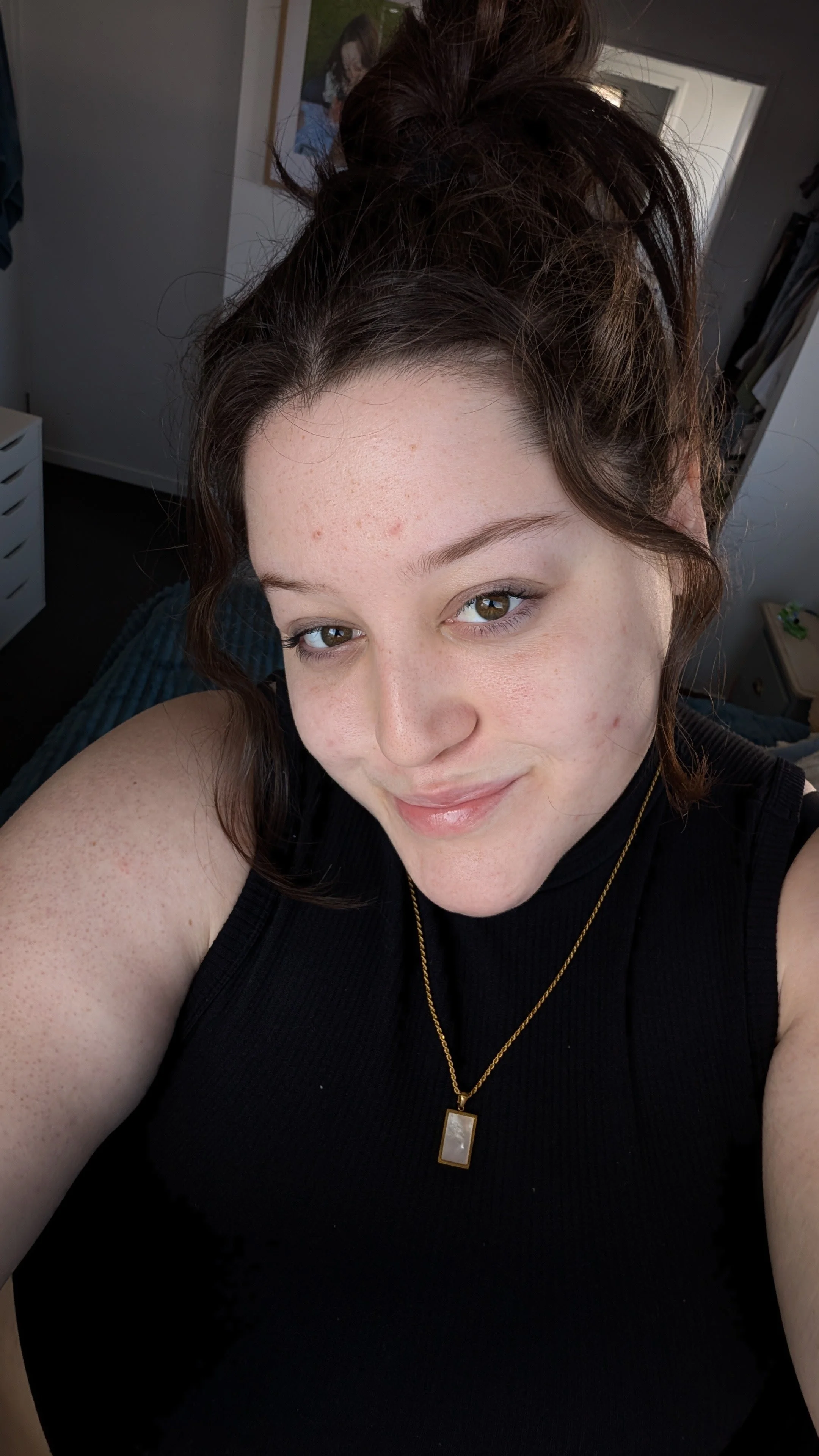A close-up selfie of a young woman with brown hair tied in a messy bun, wearing a black sleeveless top and a gold necklace with a rectangular pendant, smiling softly in a bedroom.