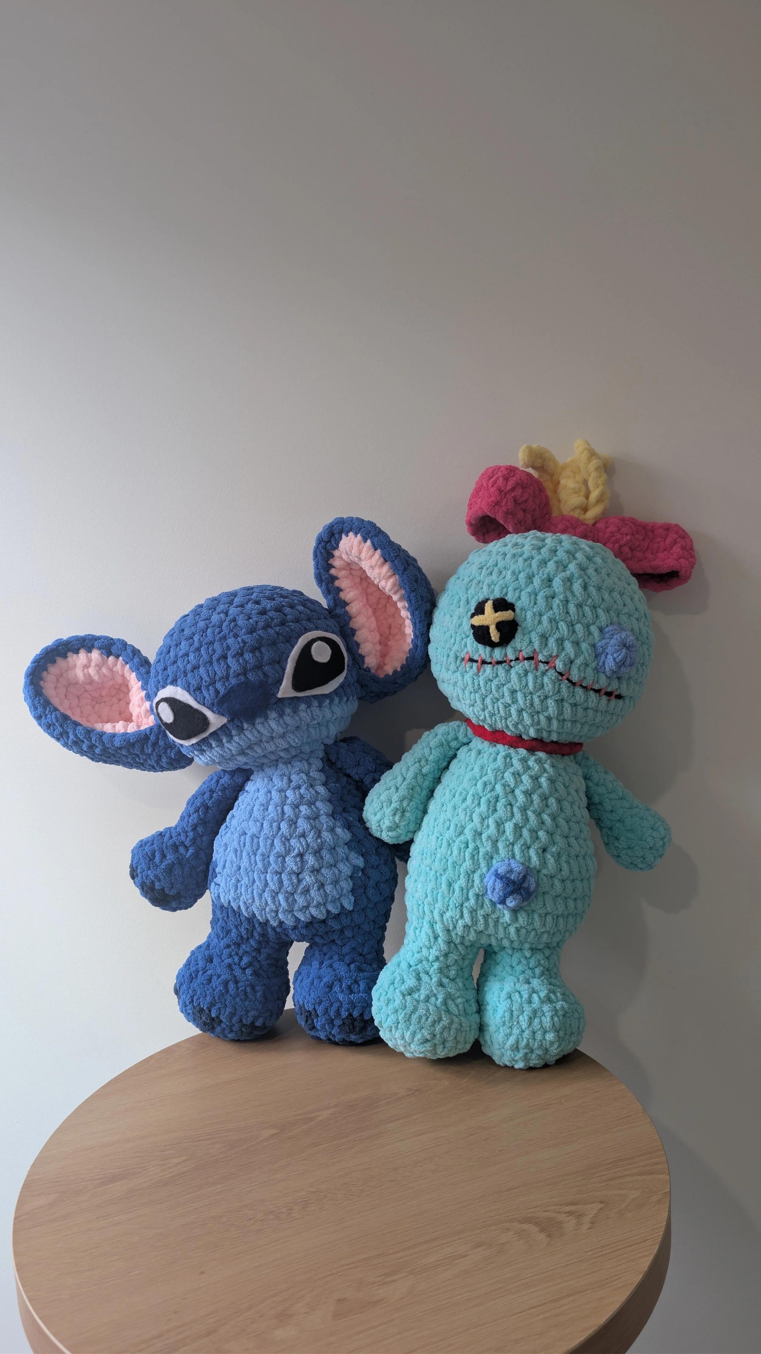 Two large crocheted stuffed animals, one blue mouse and one light blue creature with pink ears, sitting on a wooden table against a plain white wall.