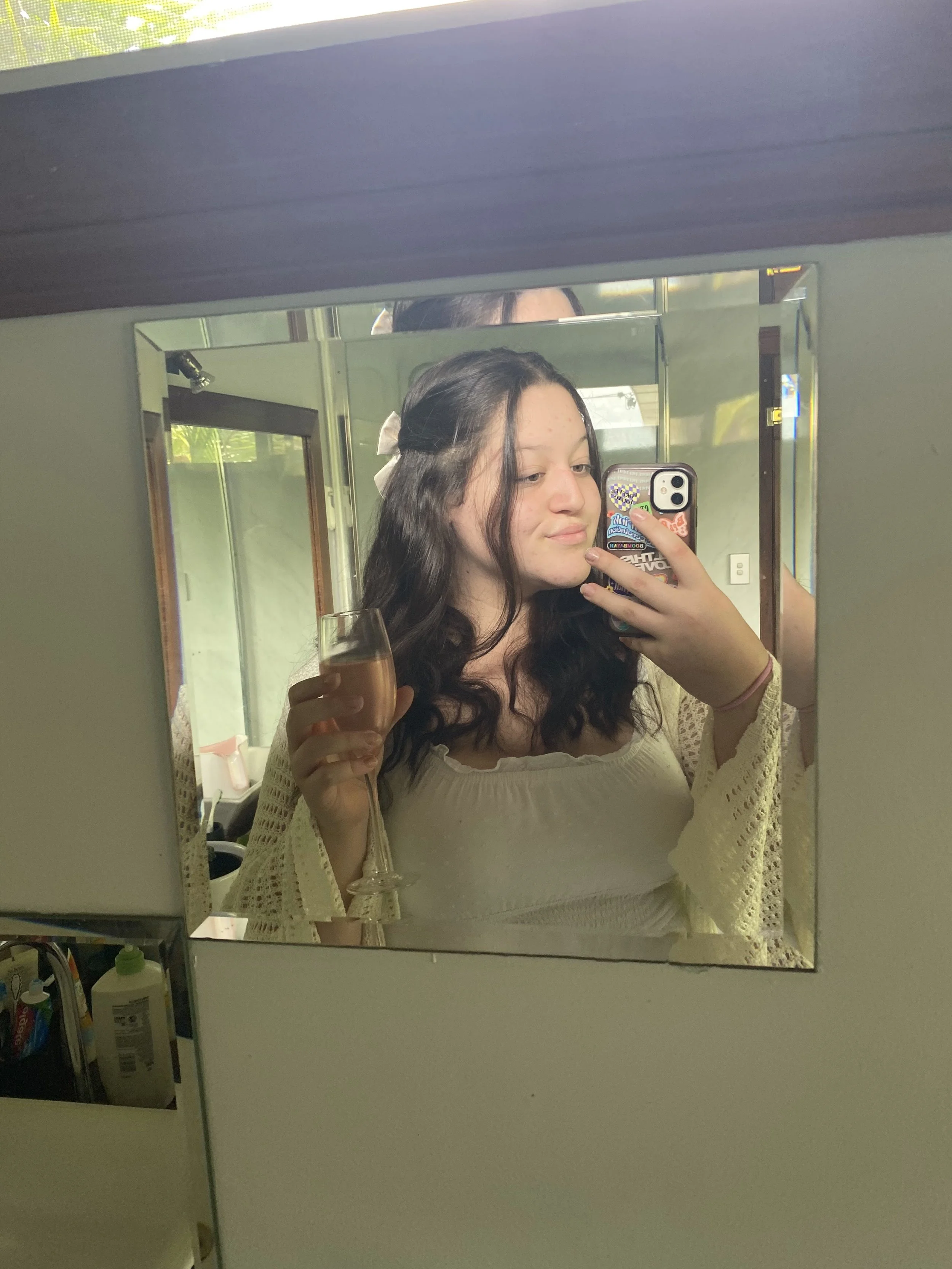 Young woman taking a mirror selfie while holding a glass of champagne, with wavy dark hair, wearing a white top and a beige cardigan.
