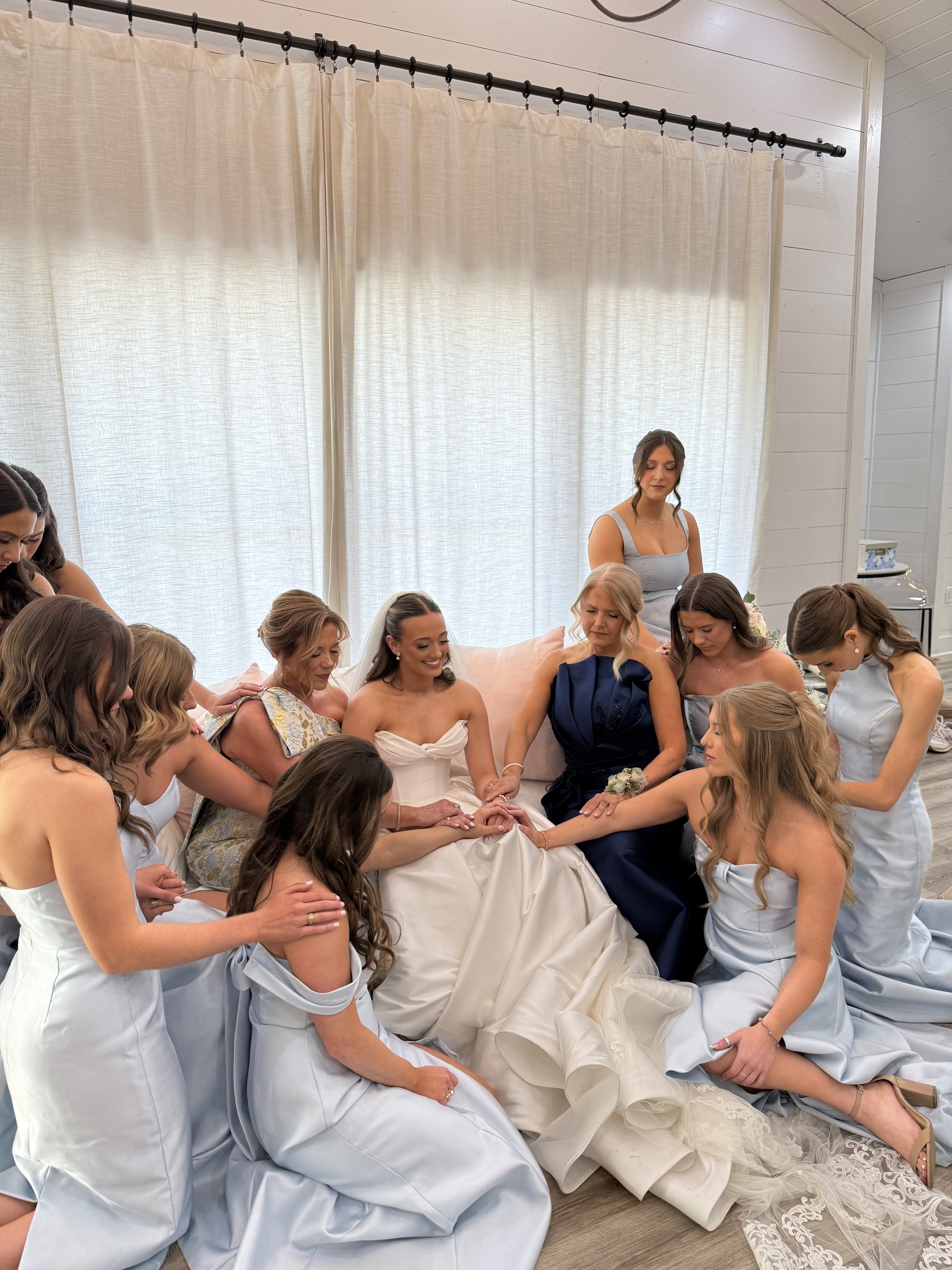 Bridal party gathered around a bride in a white wedding gown, holding her hands and praying or offering support, with bridesmaids in matching light blue dresses and a woman in a dark blue dress, inside a bright room with light-colored curtains.